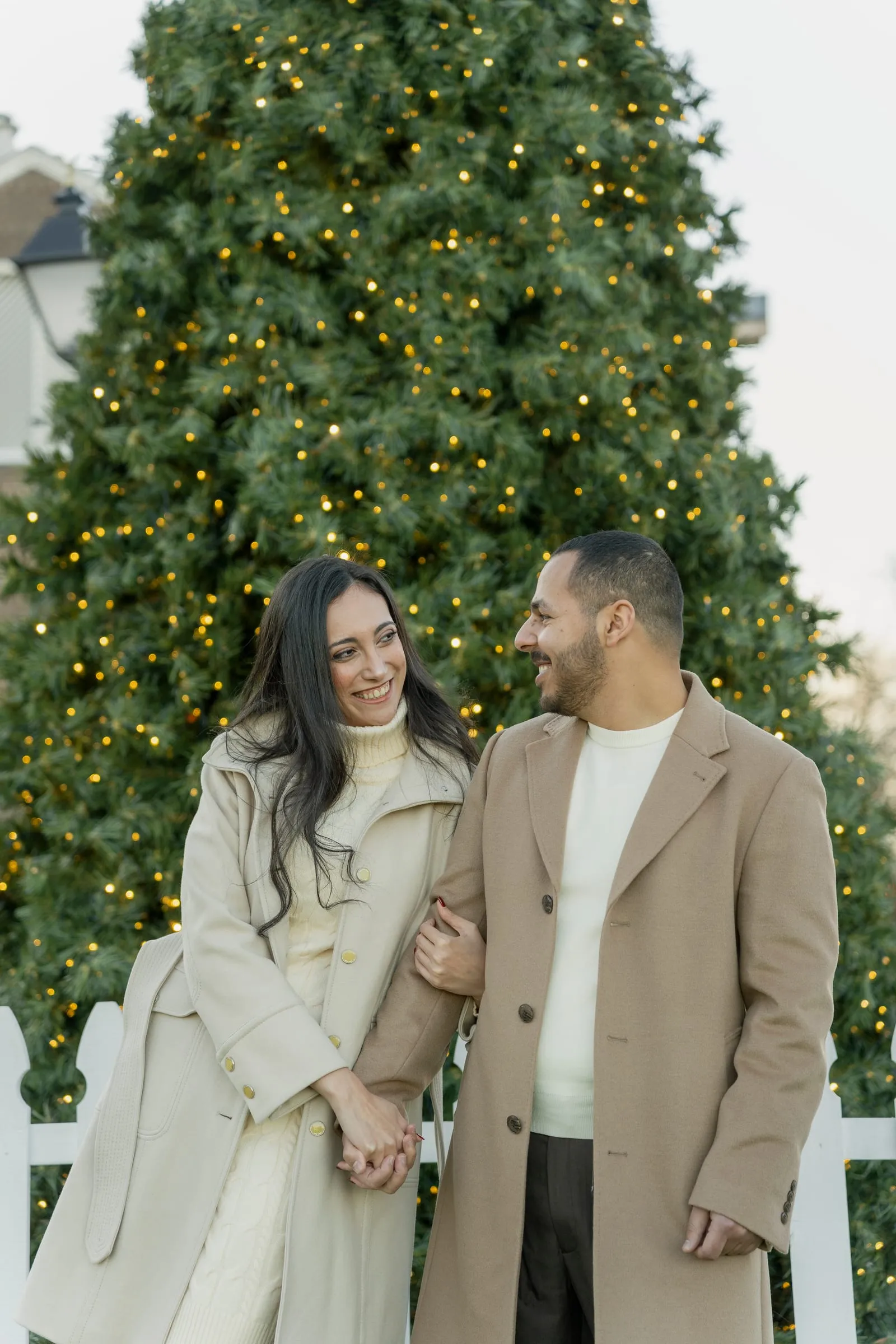 Merona smiling up at Abanoub as they stand arm in arm by the white picket fence and Christmas tree