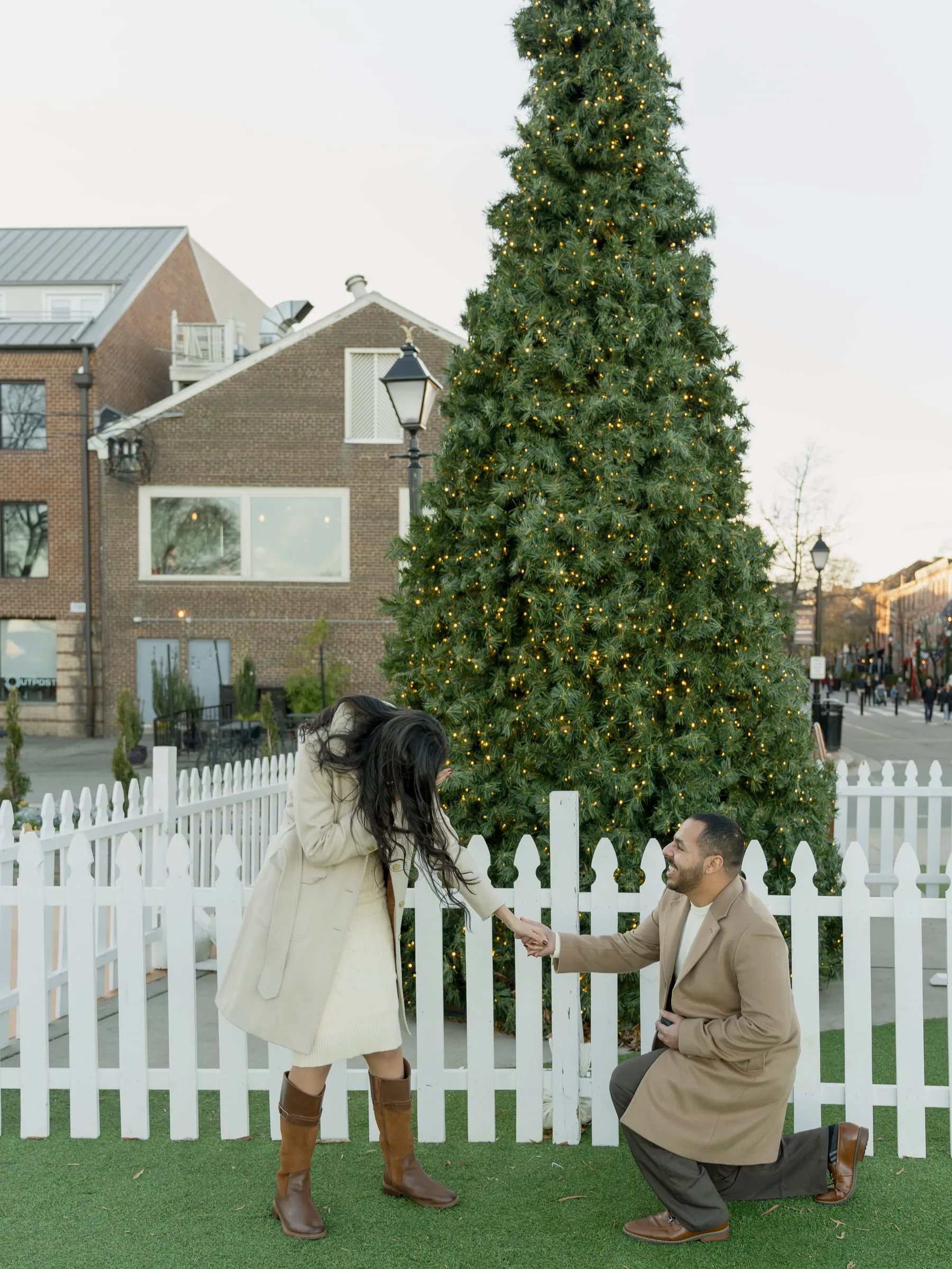 Abanoub dropping to one knee behind the white picket fence as Merona turns around by the Christmas tree