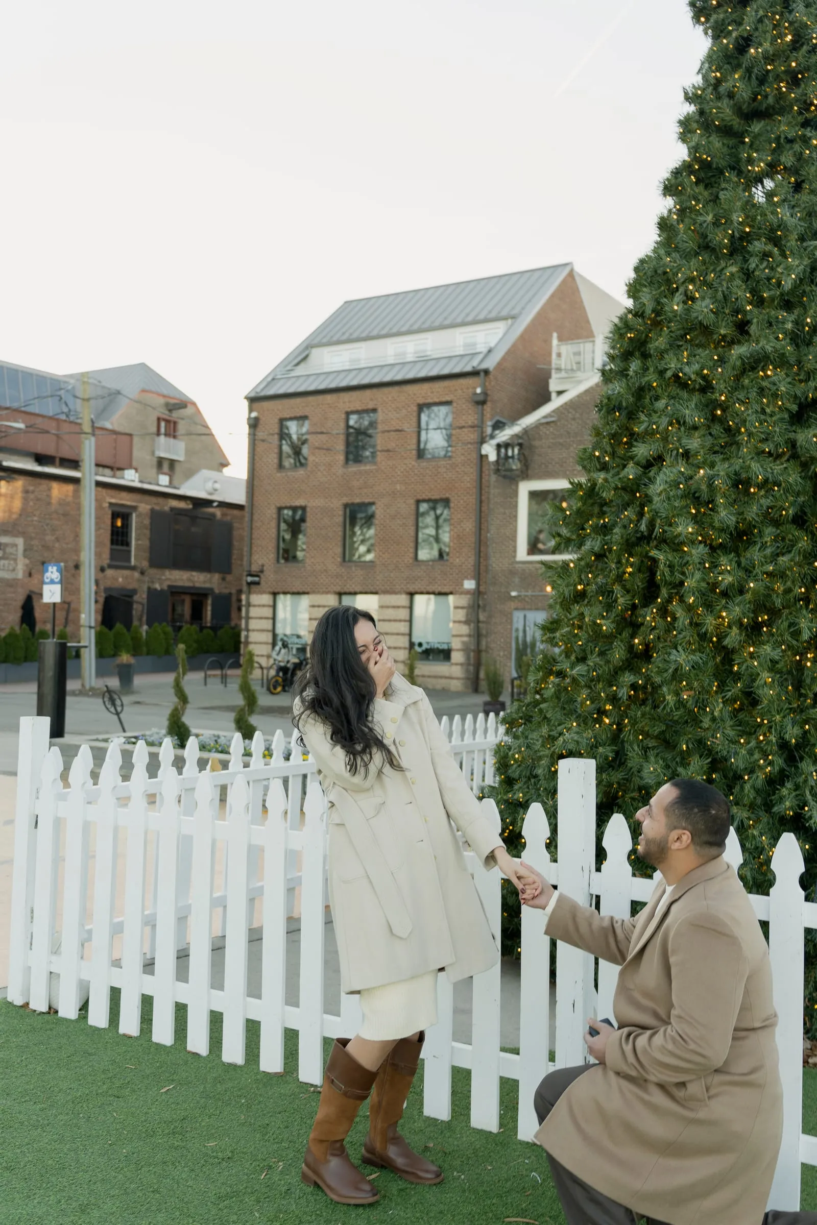 Abanoub on one knee holding Merona's hand by the picket fence, the brick buildings of Old Town behind them