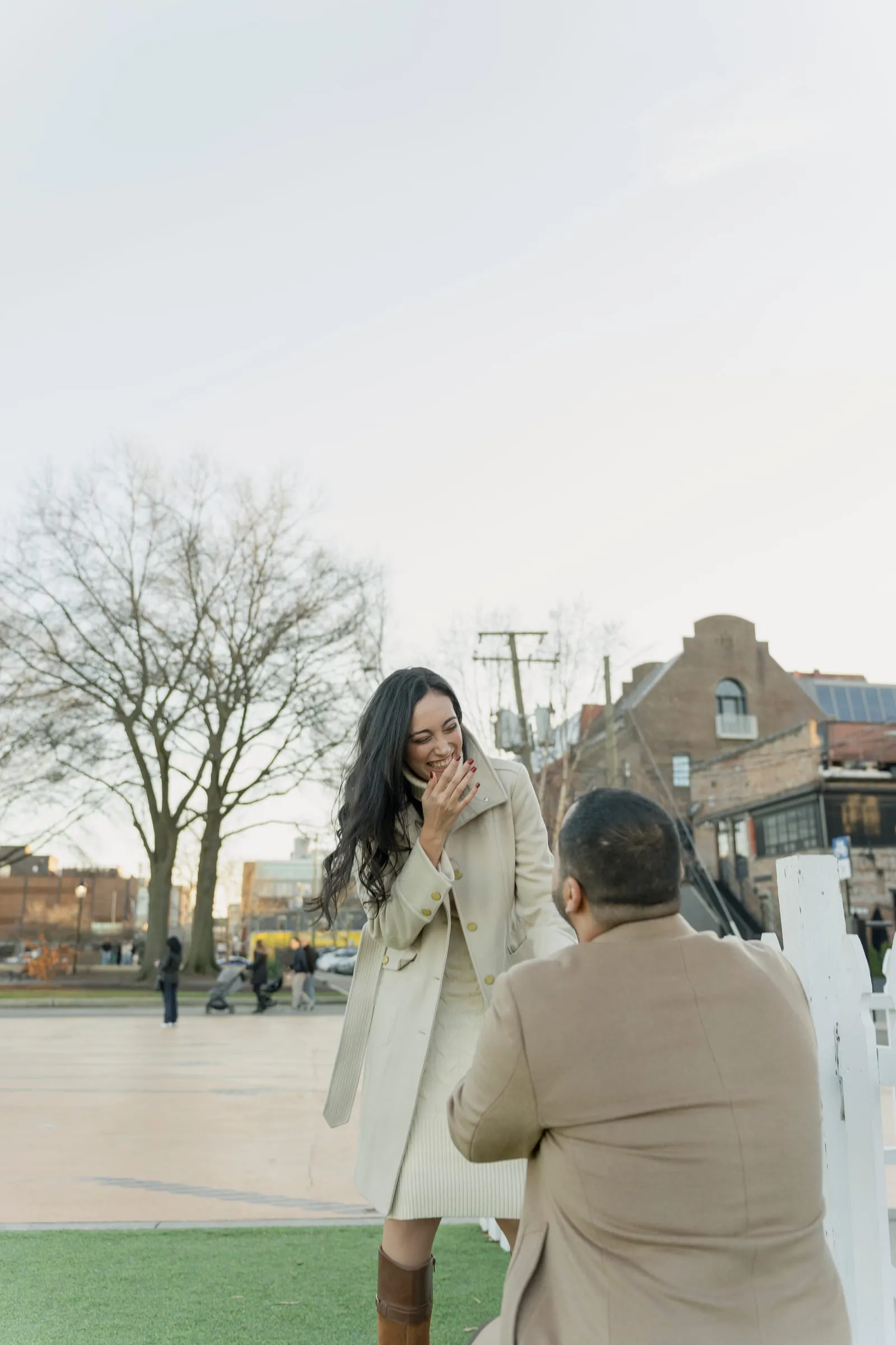 A wide view of Abanoub proposing to Merona on the lawn, bare trees and brick buildings behind them