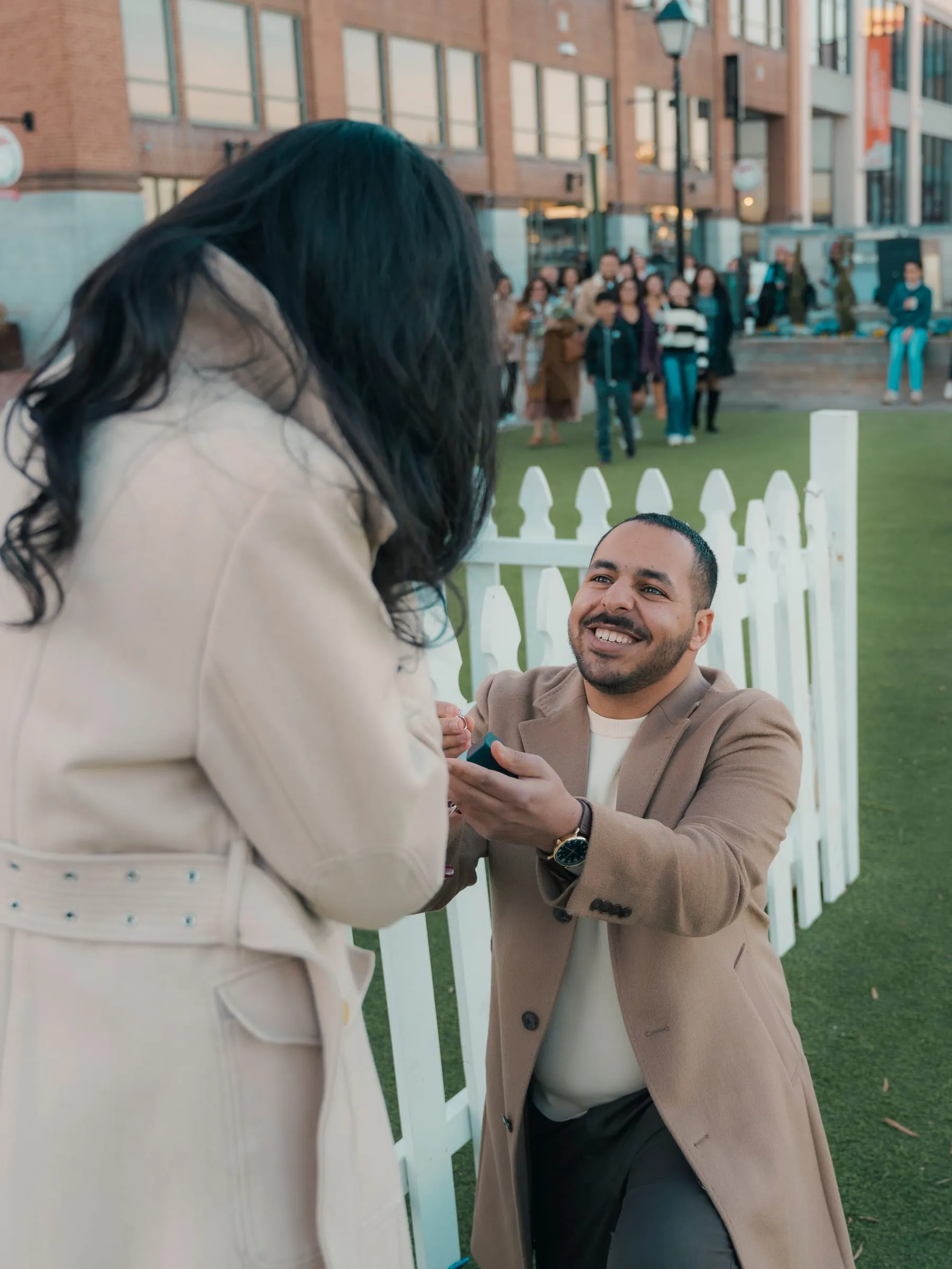 Abanoub smiling up from one knee and opening the ring box, a crowd of friends gathering behind