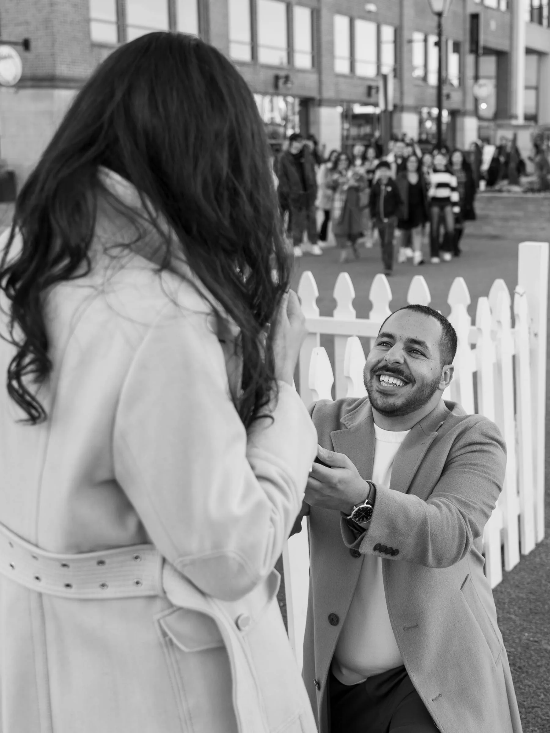 Black and white photo of Abanoub on one knee holding out the ring, friends watching in the background