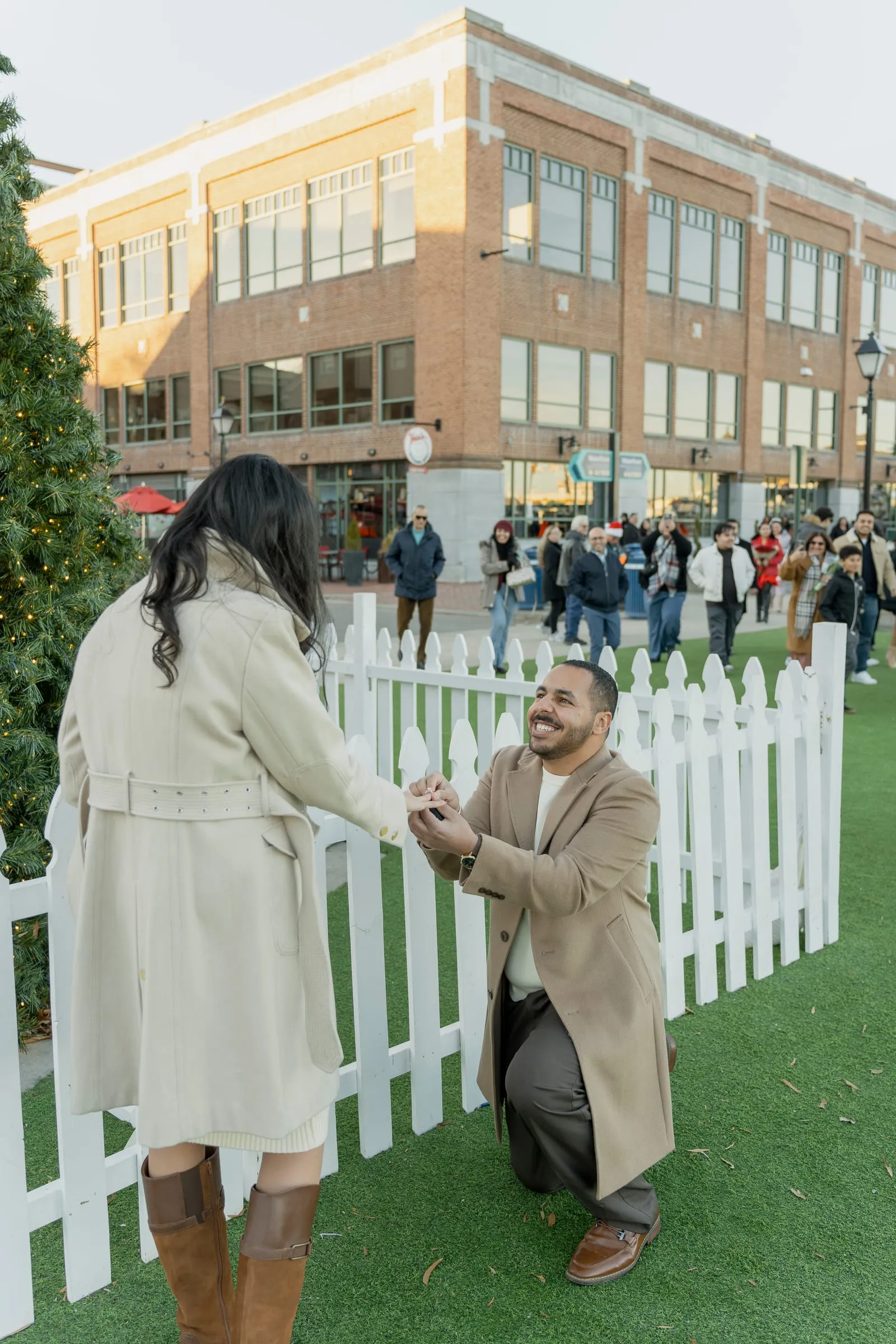 Abanoub on one knee placing the ring on Merona's hand beside the white picket fence