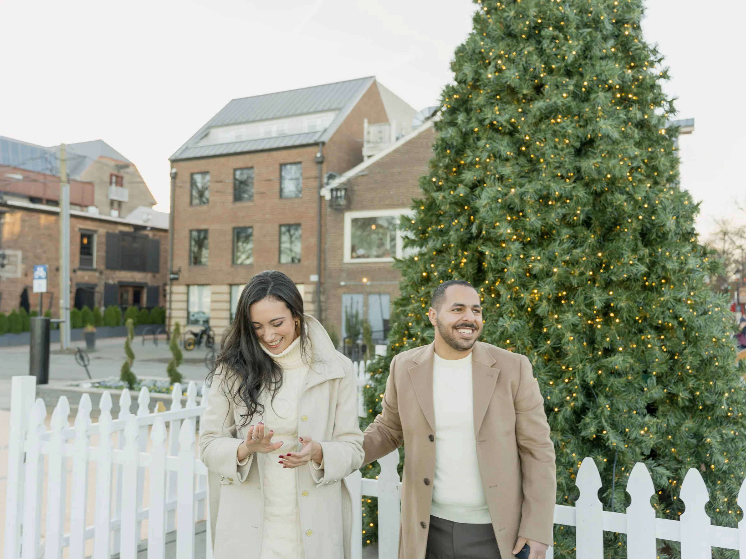Merona admiring the engagement ring while Abanoub laughs, the tree and brick buildings behind them