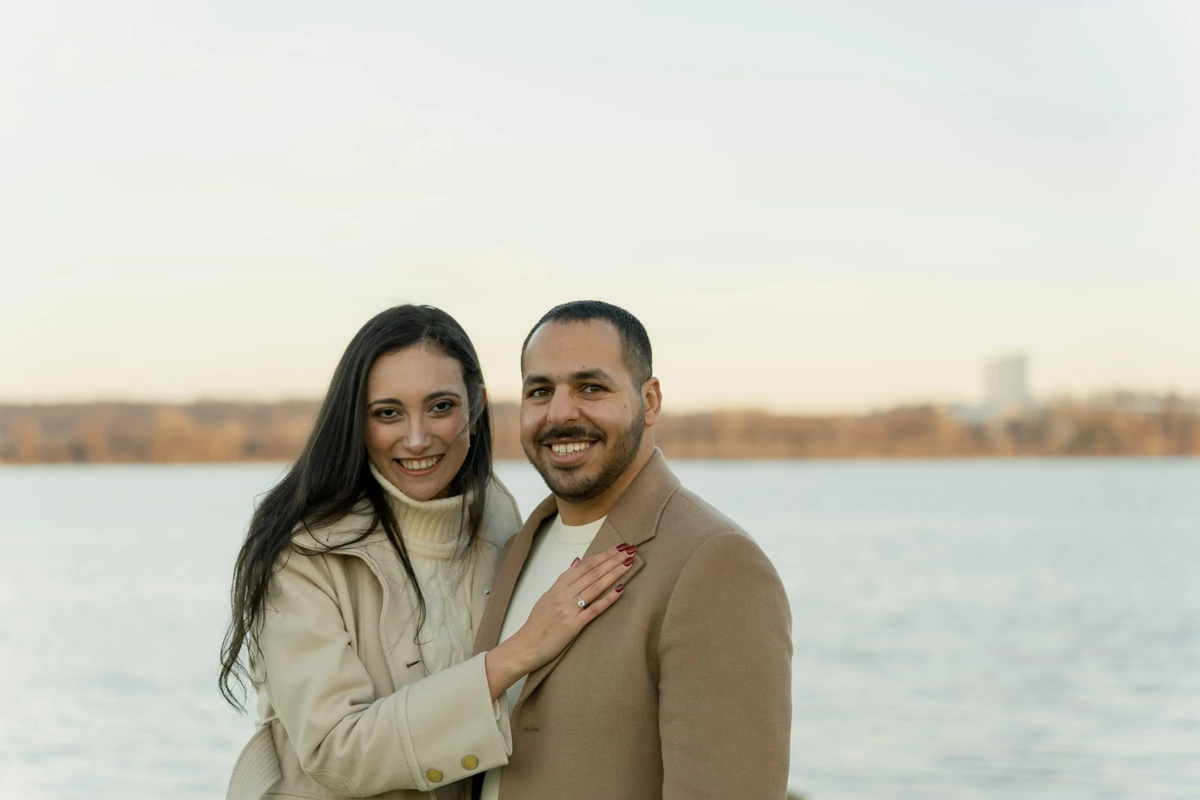 Abanoub and Merona smiling together by the water, Merona's ring showing against Abanoub's coat
