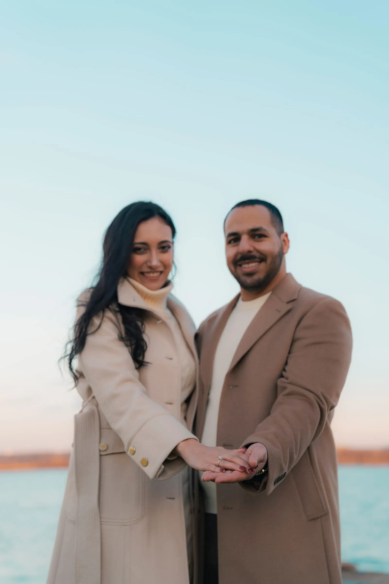 Abanoub and Merona smiling side by side against a blue sky, holding hands to show the ring