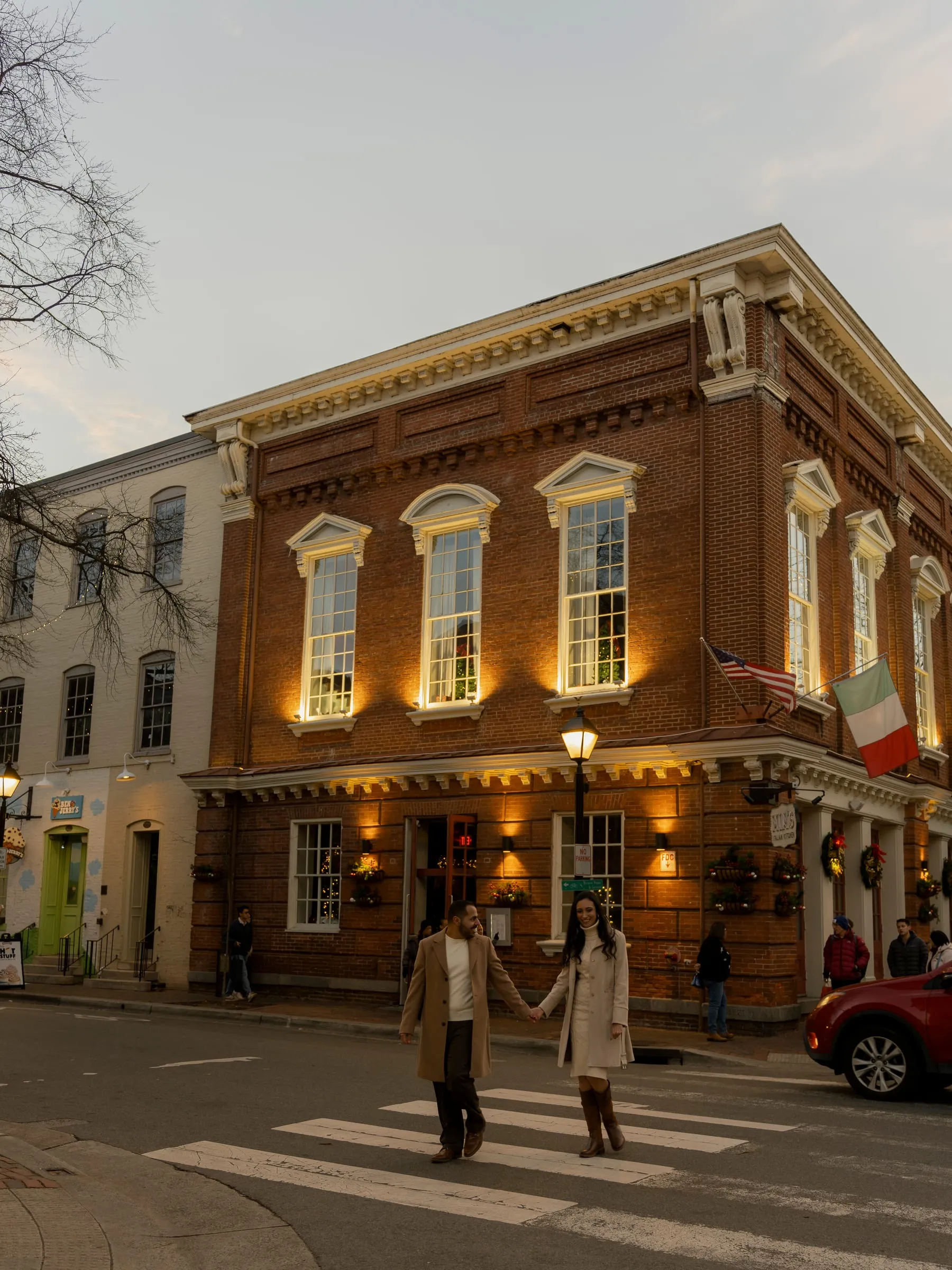 A wide view of Abanoub and Merona crossing the street in front of a lit brick building at dusk