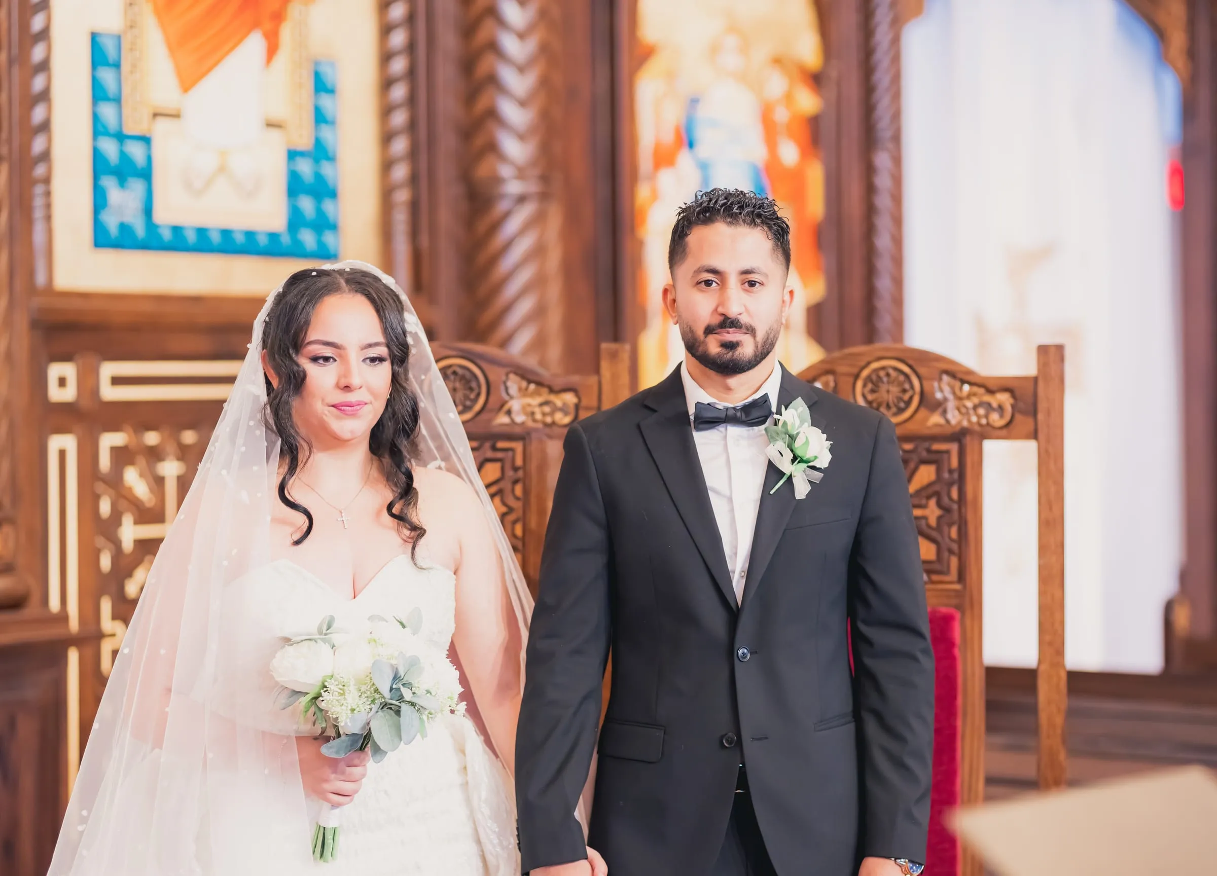 Gina holding a white bouquet beside Fady at the altar, painted icons behind them