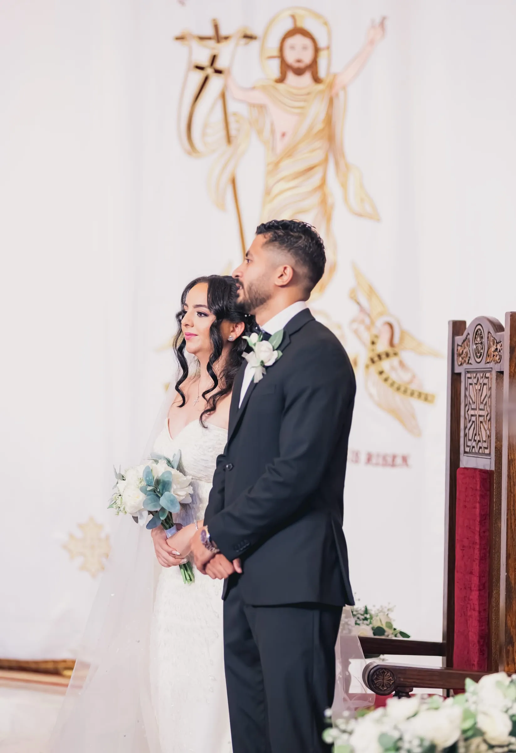 Gina and Fady standing before a white backdrop embroidered with the risen Christ