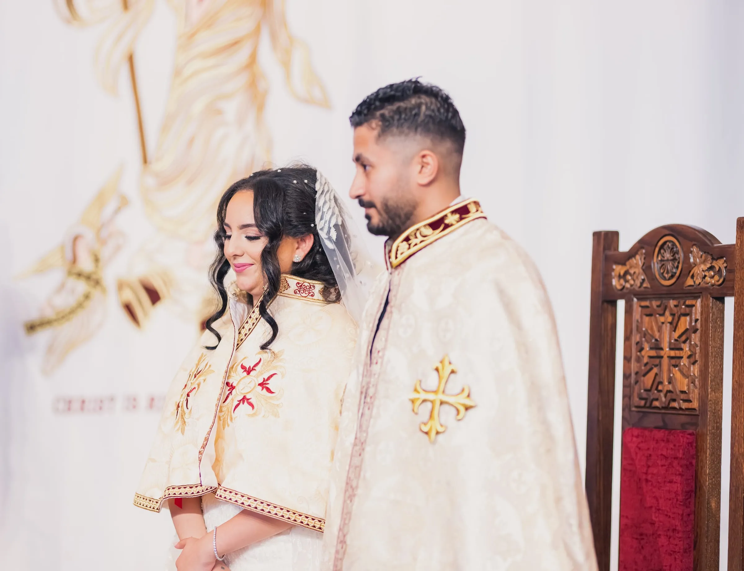Gina and Fady in embroidered ceremonial robes, smiling beside a carved wooden throne