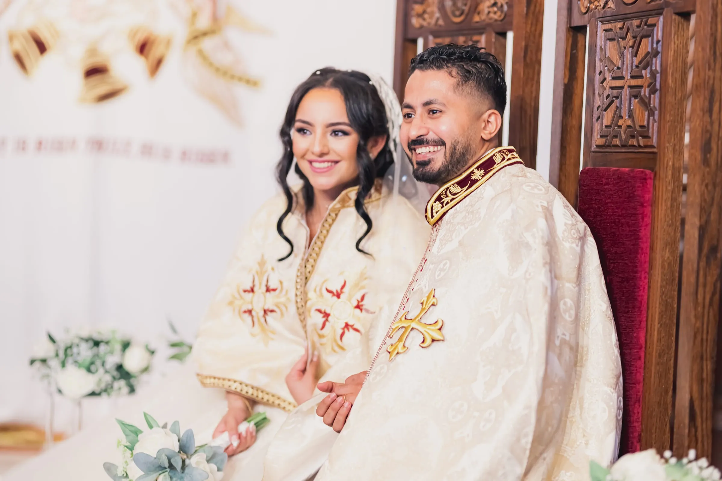 Gina and Fady smiling on the wedding thrones, white flowers at their feet