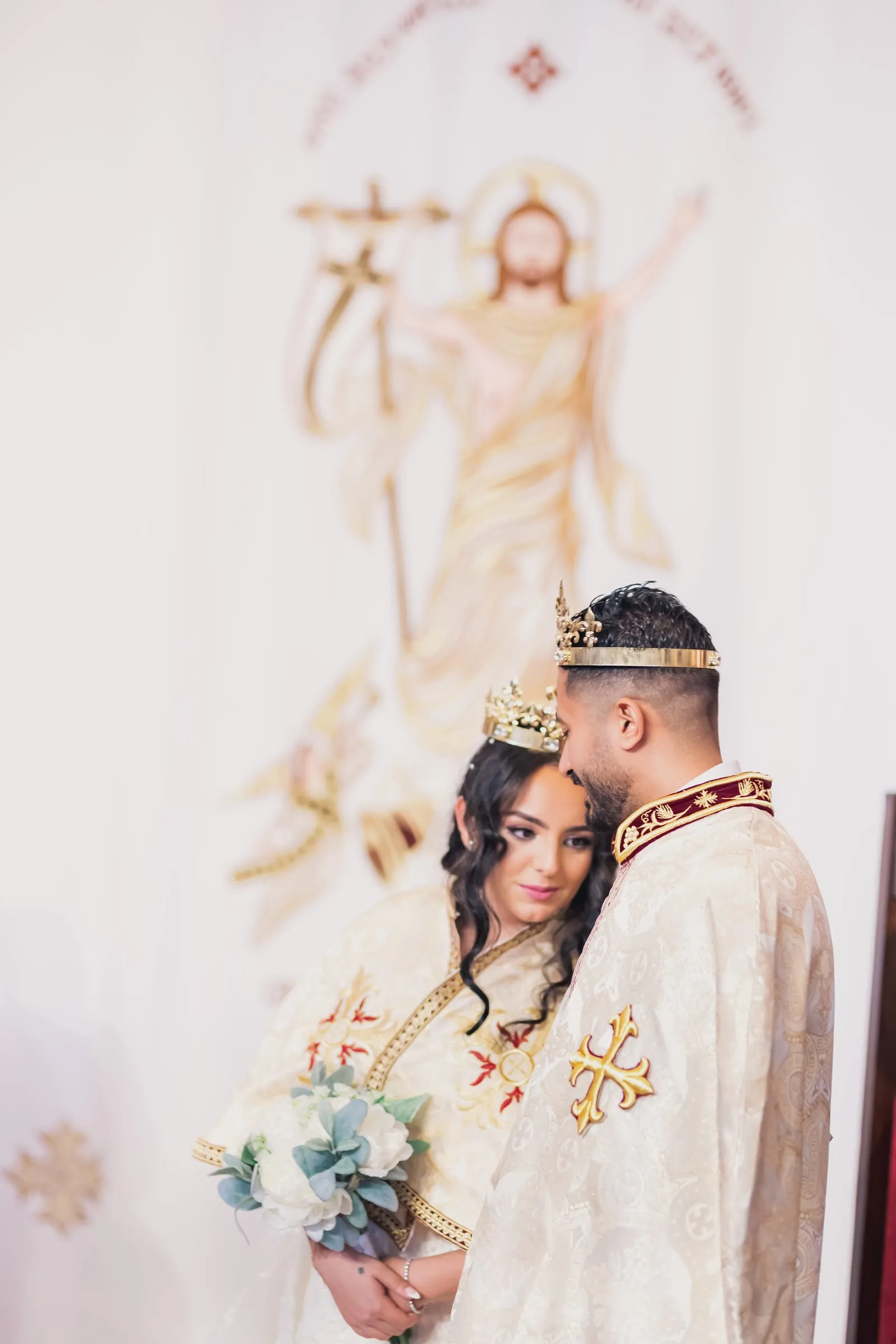 Gina resting on Fady's shoulder, both crowned, before the embroidered backdrop