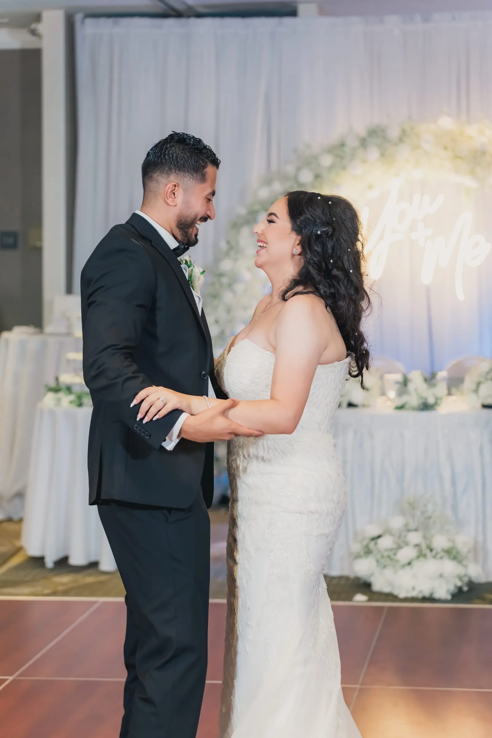 Gina and Fady sharing their first dance in front of a white floral arch and a glowing sign