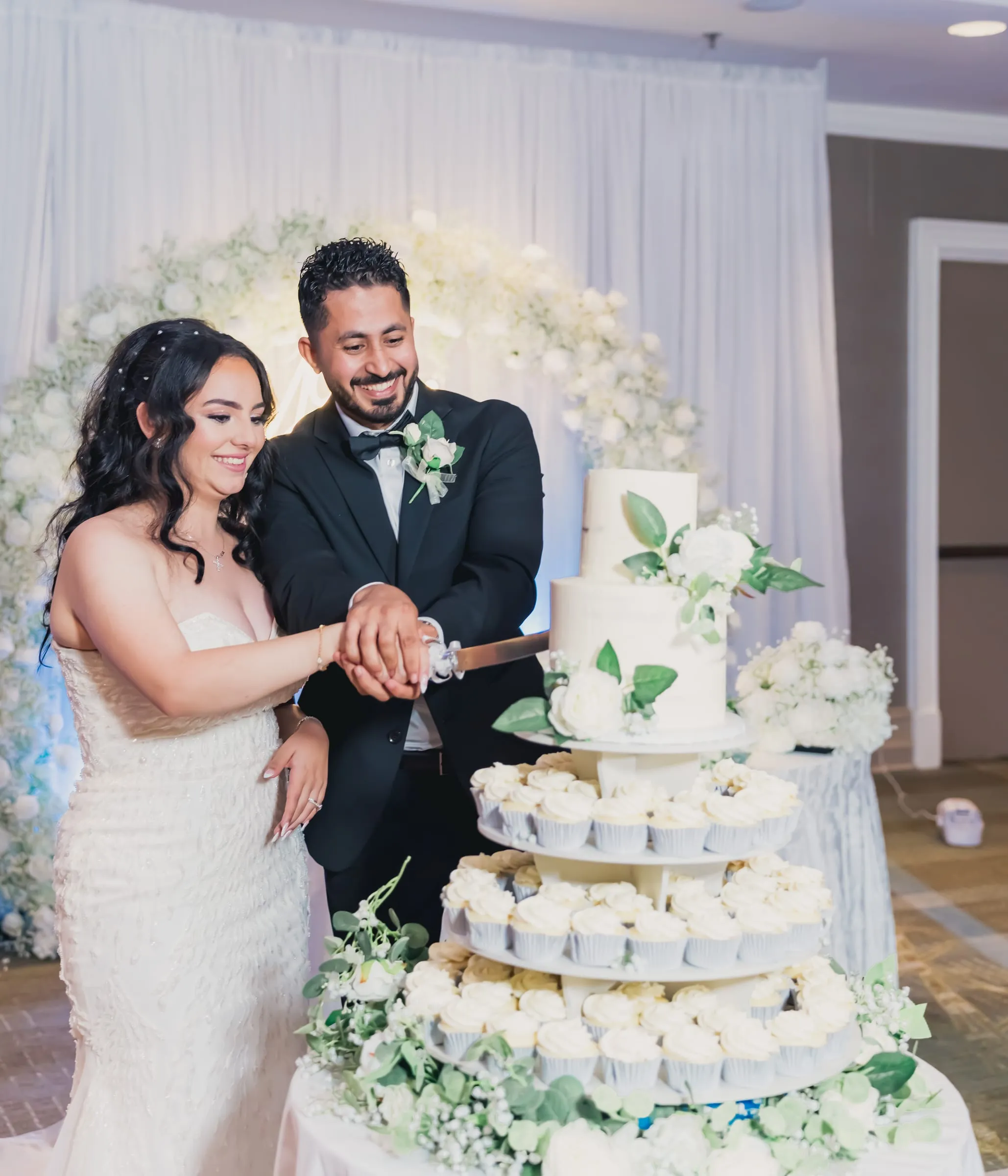 Gina and Fady cutting their tiered cake and cupcake tower in front of a white floral arch