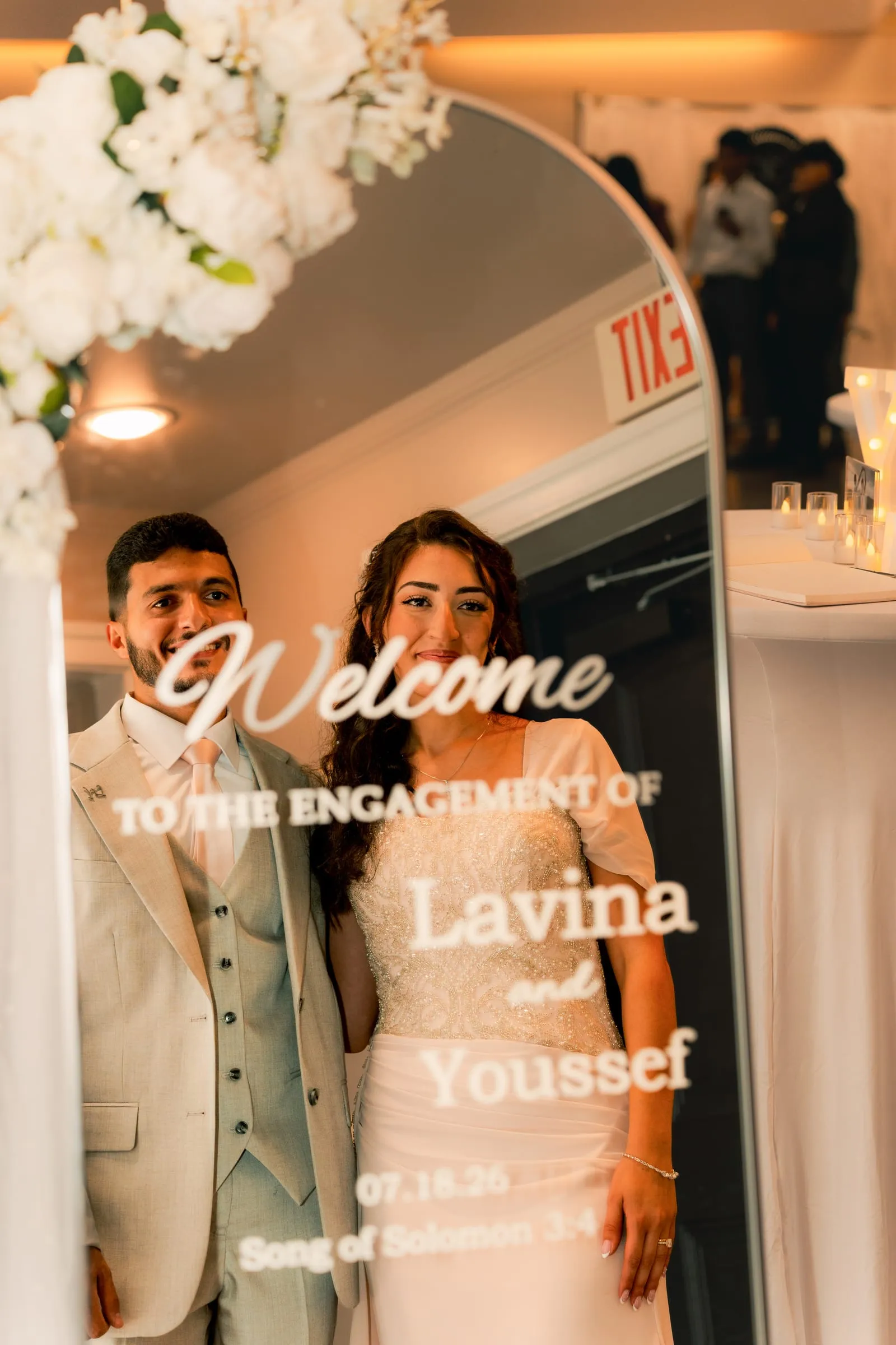 Lavina and Youssef smiling behind an engraved mirror welcoming guests to their engagement celebration