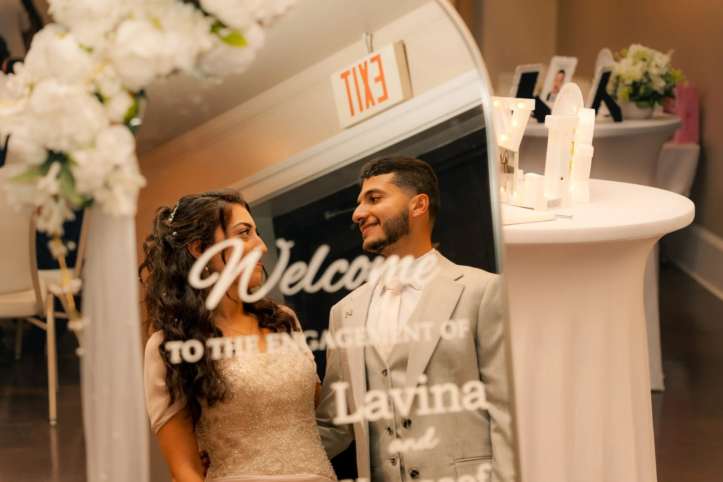 Lavina and Youssef smiling at each other, seen through the mirrored sign welcoming guests to their engagement