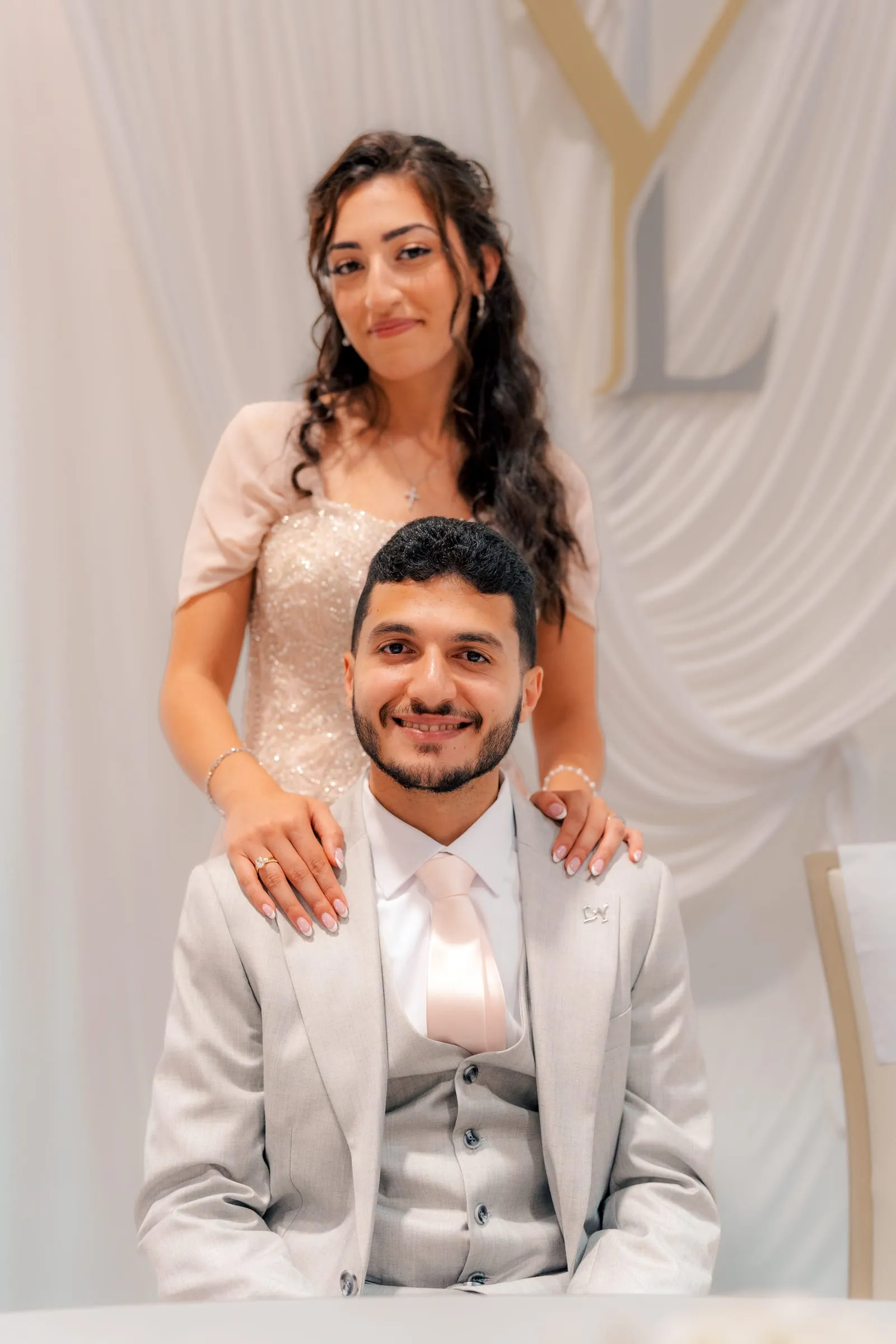 Lavina standing behind Youssef with her hands on his shoulders in front of gold Y and L monogram letters