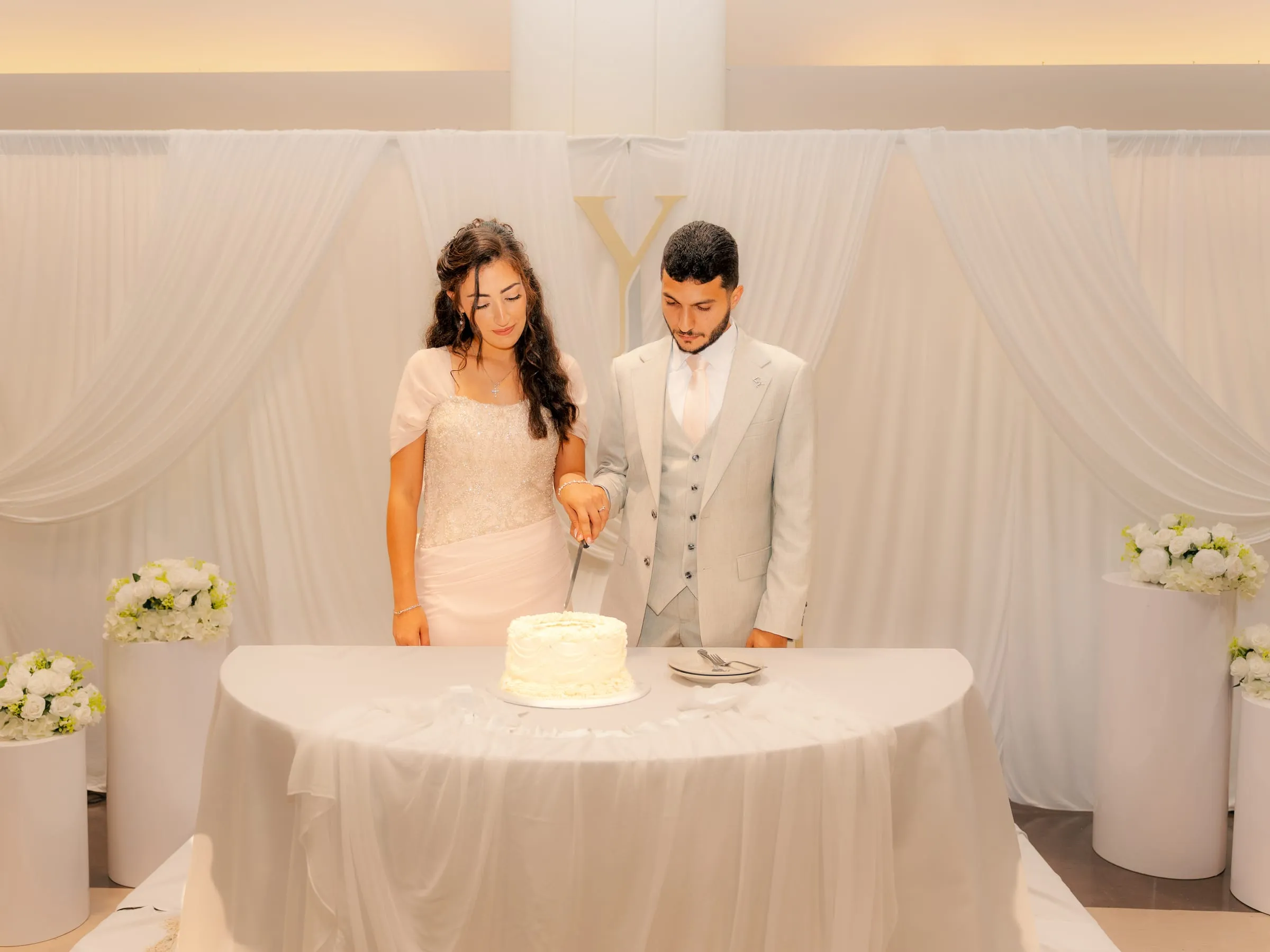 Lavina and Youssef cutting their engagement cake together in front of a white draped backdrop
