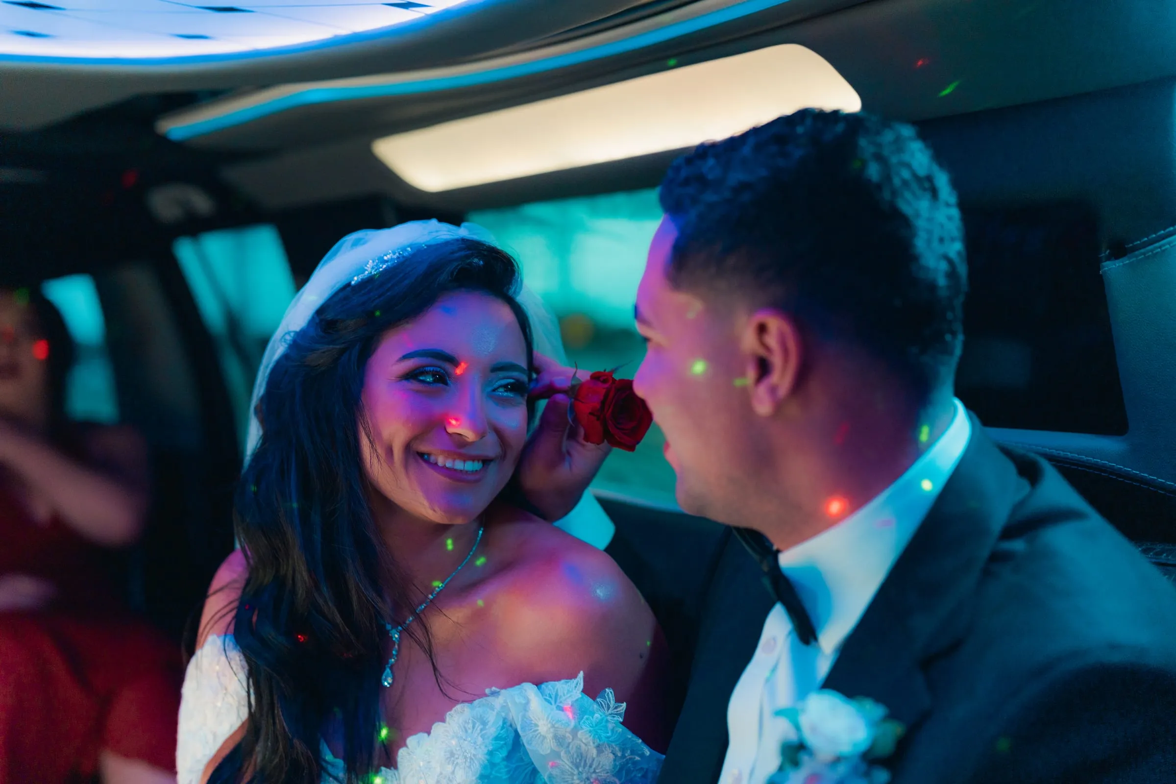 Mariam smiling with a rose held to her cheek as Mina leans in close inside the limo