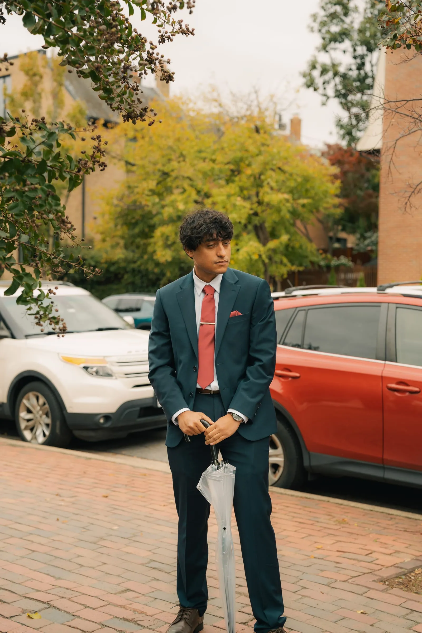 A groomsman in a navy suit and red tie standing on a brick sidewalk with an umbrella