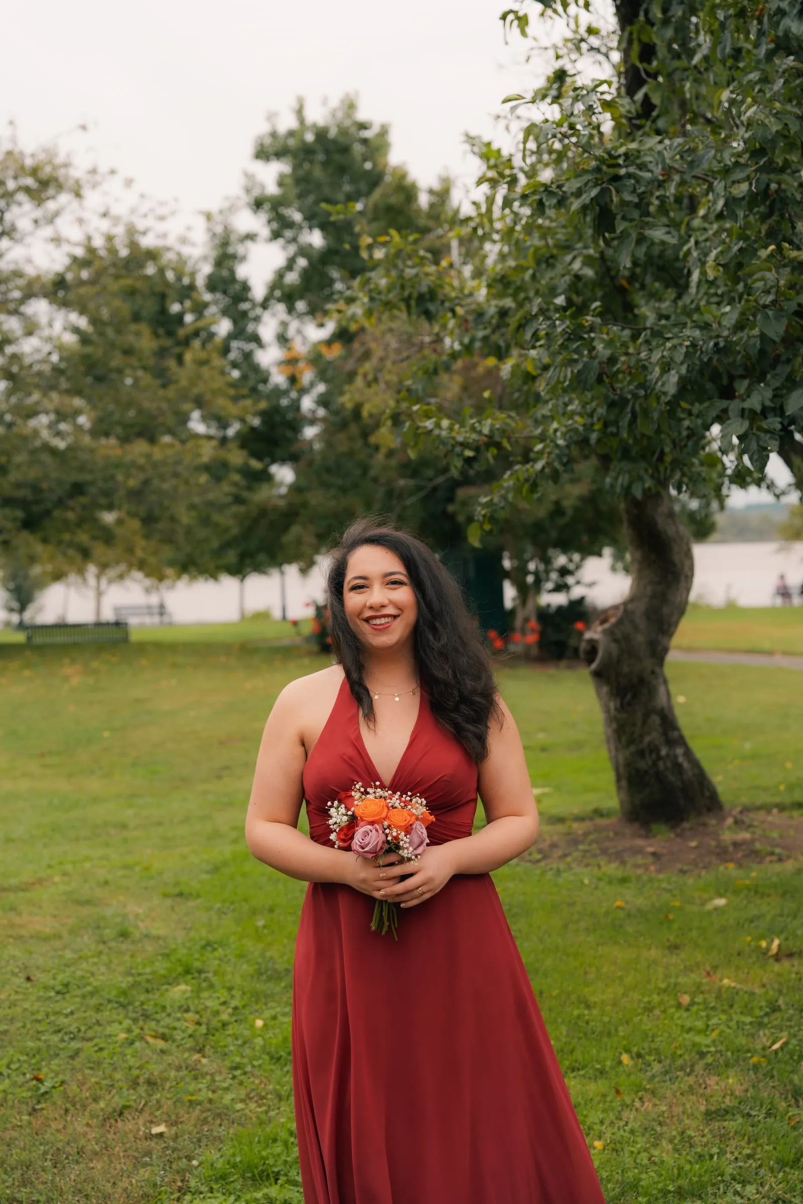 A bridesmaid in a red dress smiling with her bouquet in the park