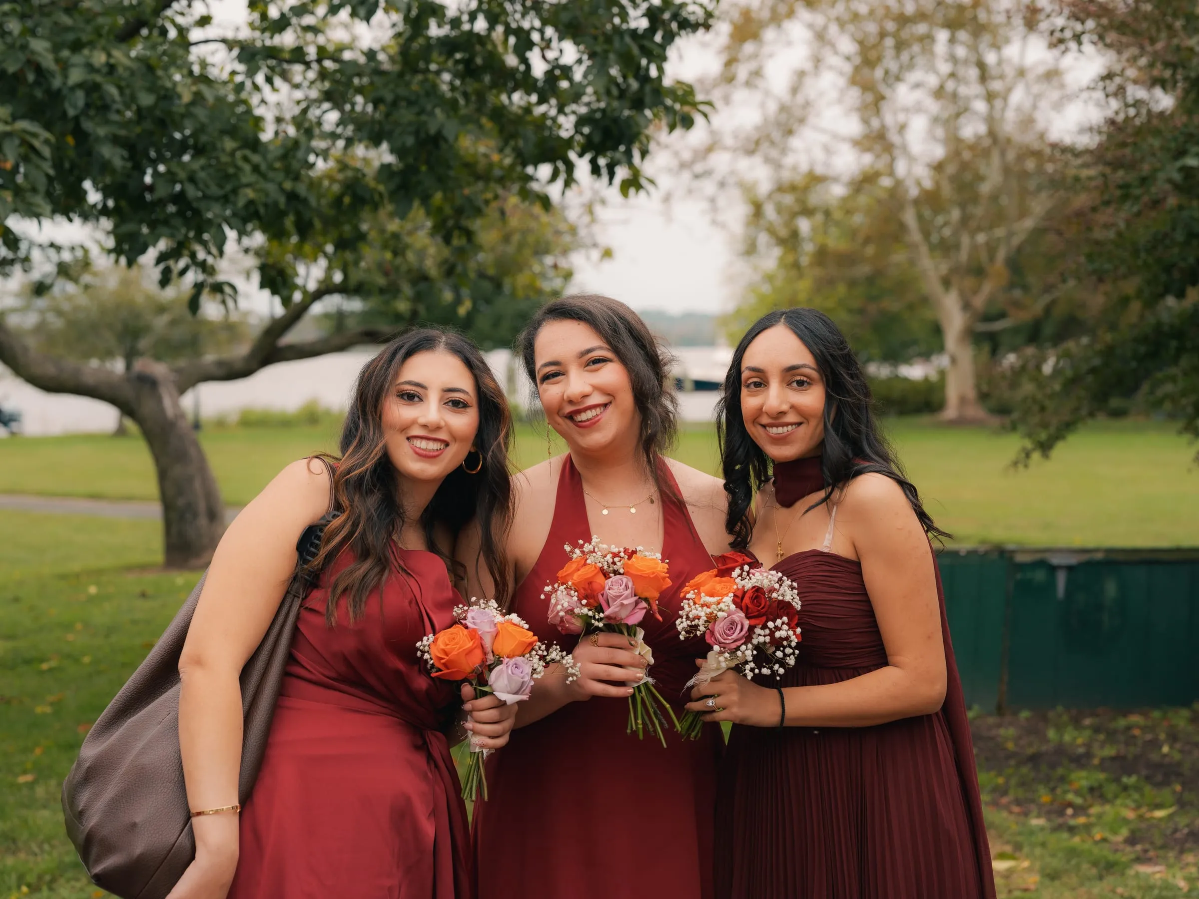 Three bridesmaids in red and burgundy dresses smiling together beside the water