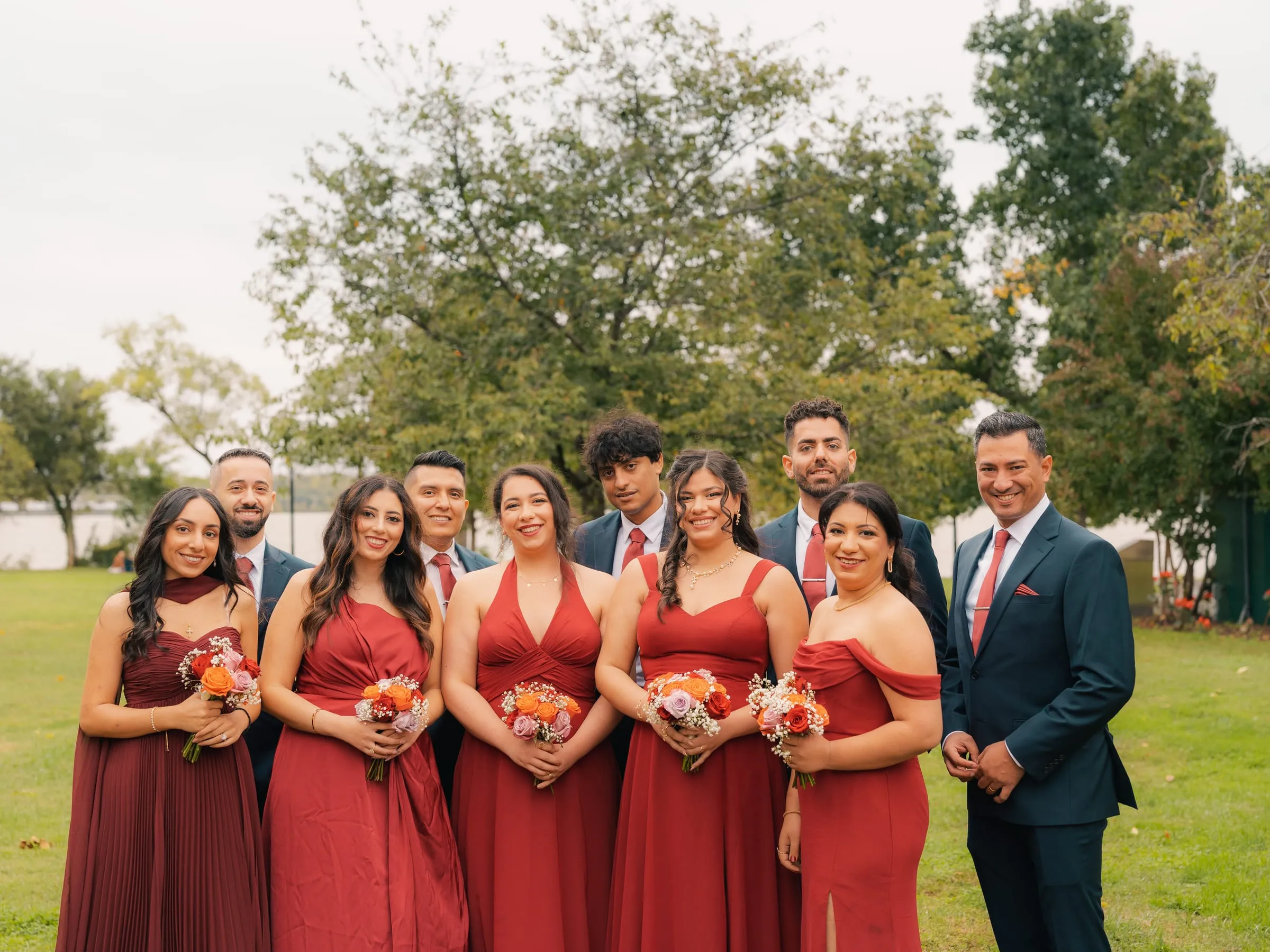 The full wedding party, bridesmaids in red and groomsmen in teal, posing together in the park