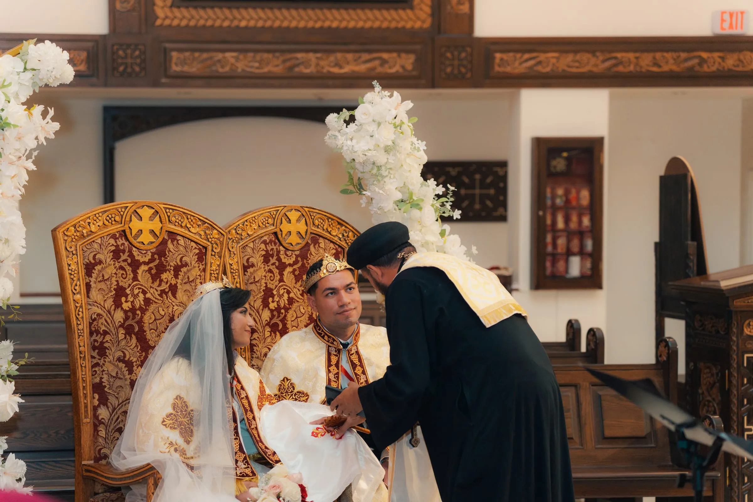 The priest blessing Mariam and Mina as they sit crowned on the wedding thrones