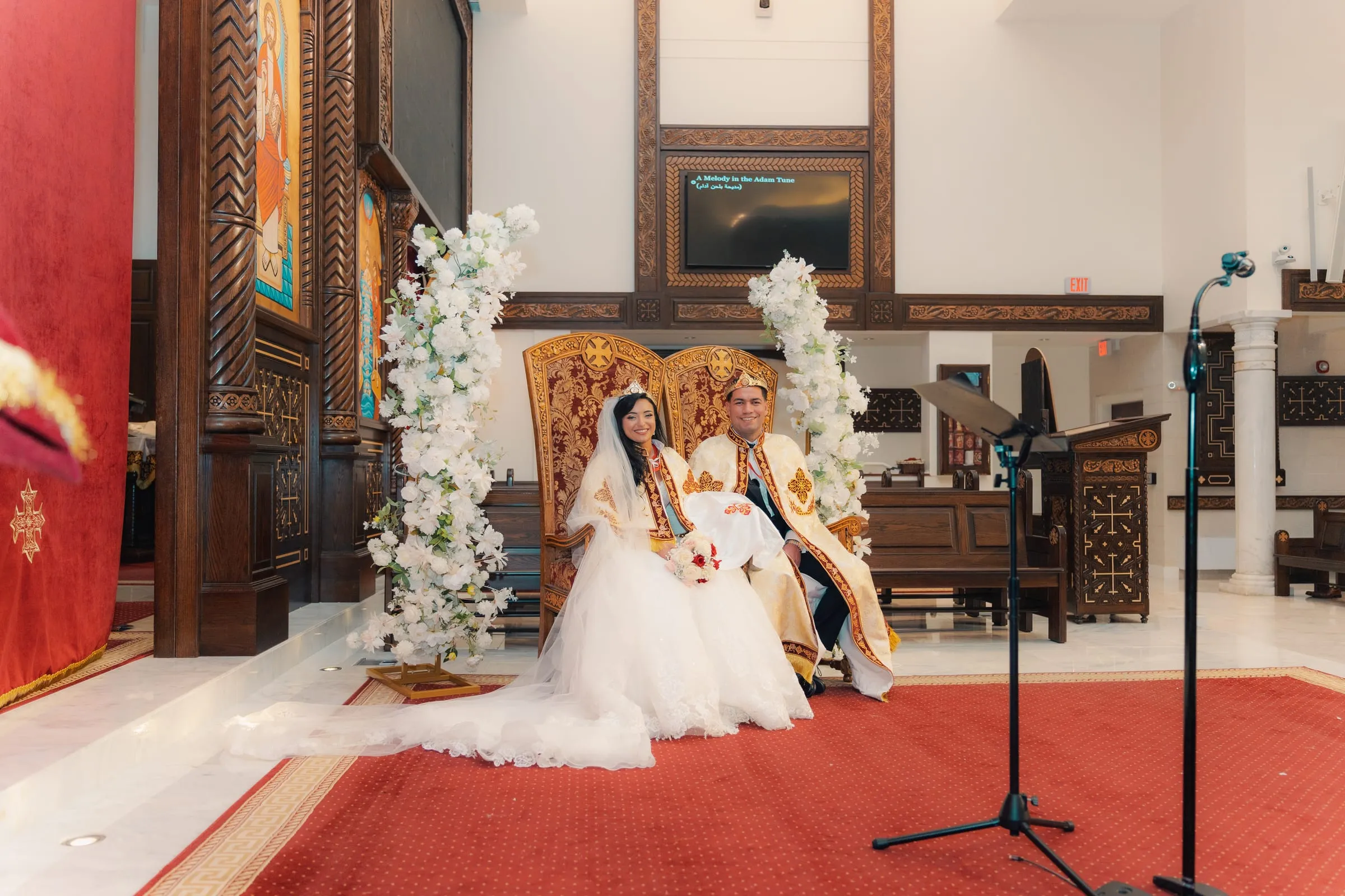 Mariam and Mina seated together beneath white floral arrangements during the crowning ceremony