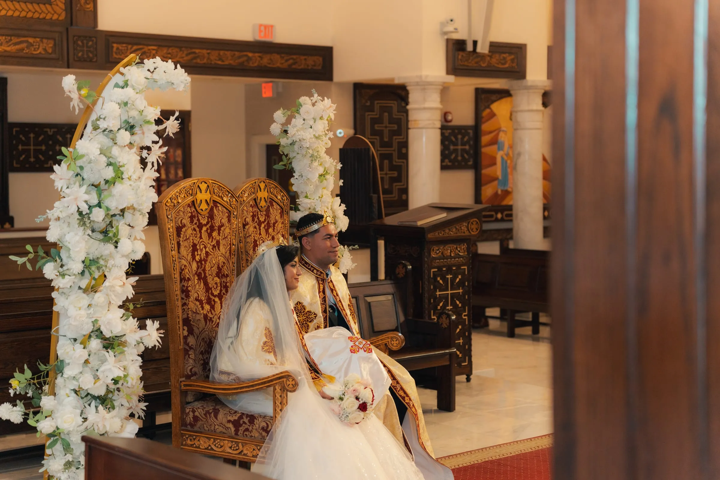 Mariam and Mina seated side by side on the wedding thrones, framed by white flowers
