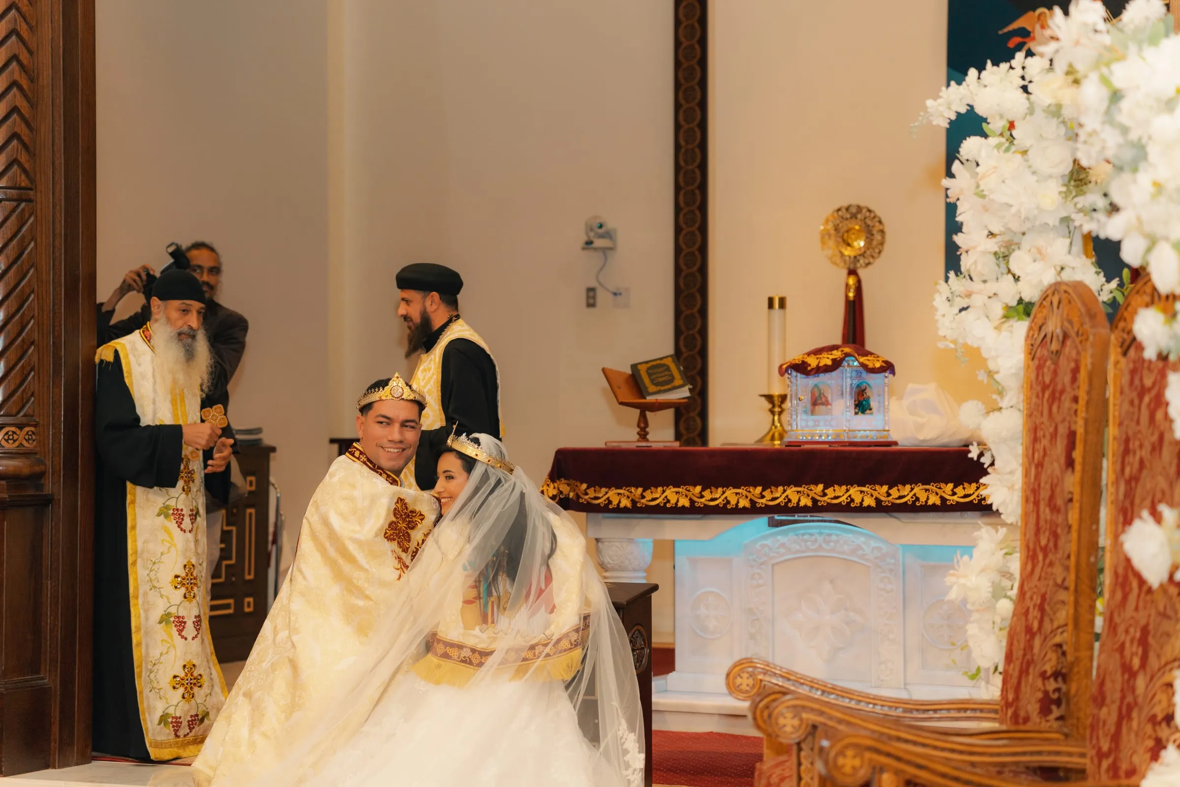 Mariam and Mina, crowned and wrapped in ceremonial robes, smiling together beside the altar