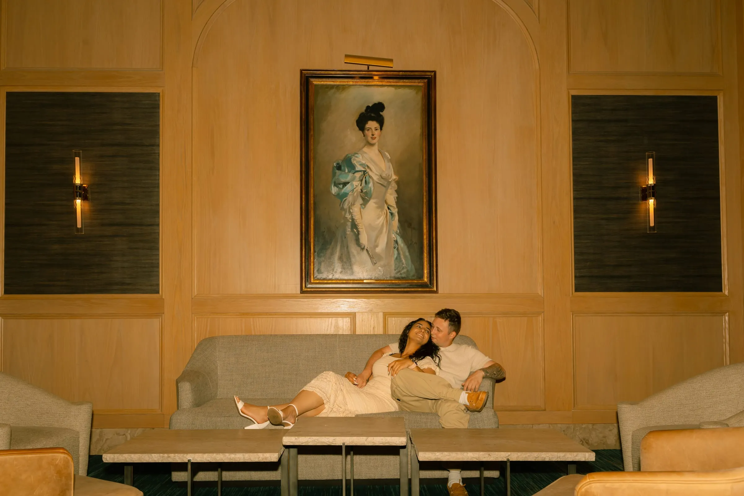 Wide view of Mariam and Thomas on the hotel lounge sofa, flanked by armchairs