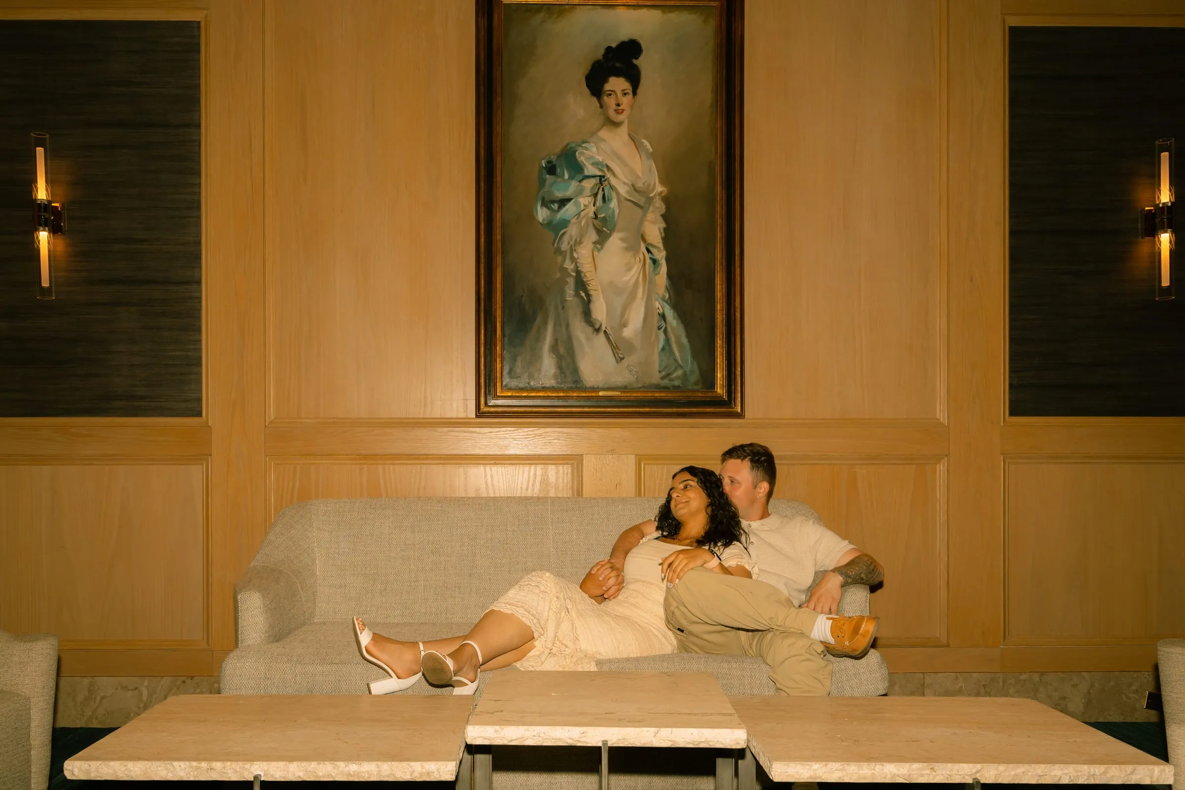 Mariam resting her head on Thomas's shoulder as they lounge on the sofa beneath the painting