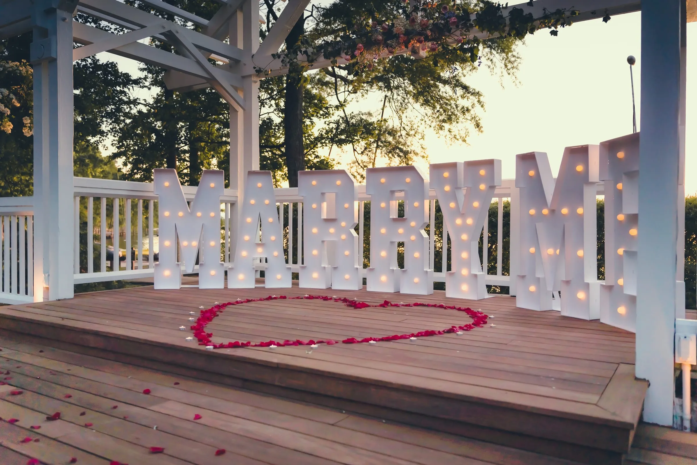 The proposal setup at sunset, marquee letters spelling MARRY ME behind a rose petal heart