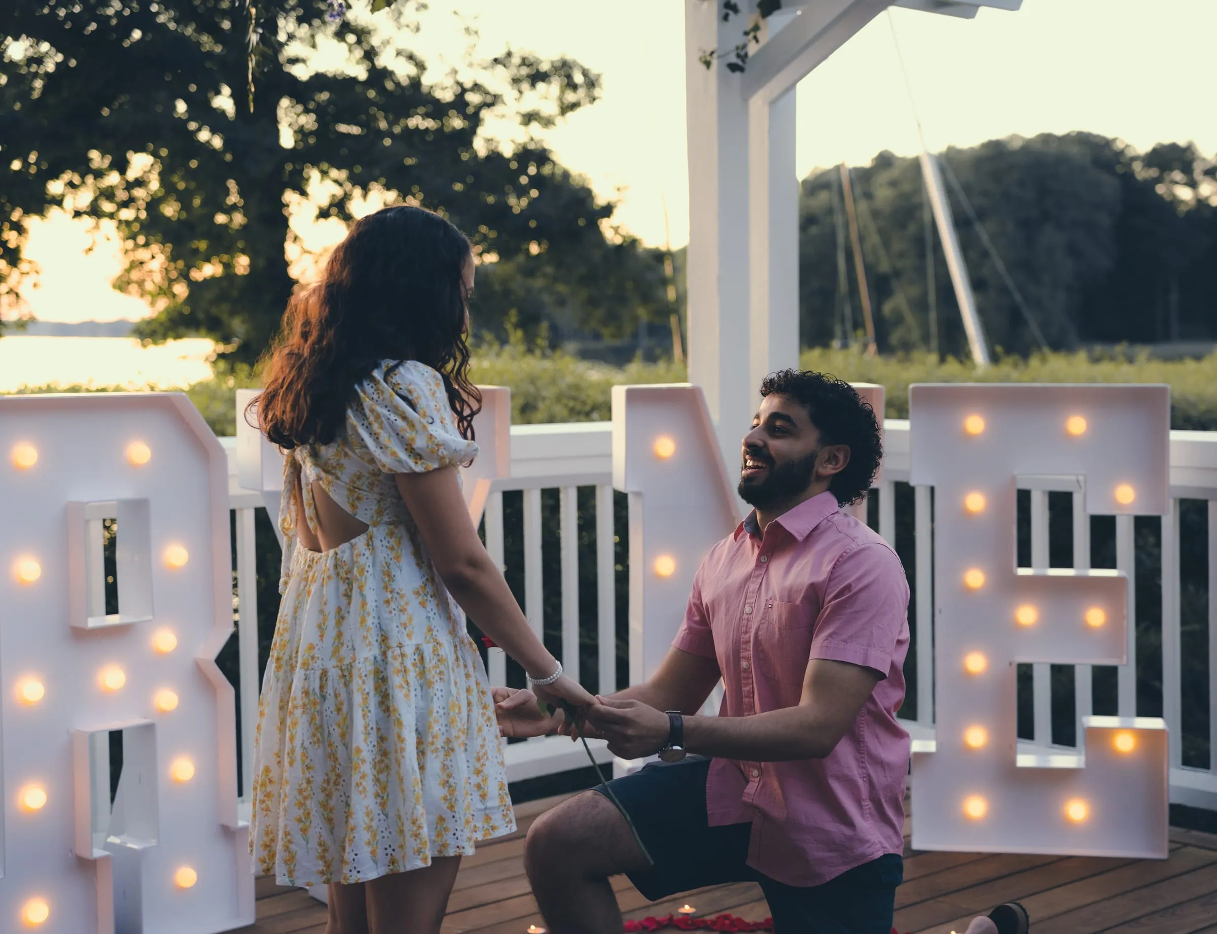 George on one knee holding Marina's hands in front of the glowing marquee letters, the water behind them