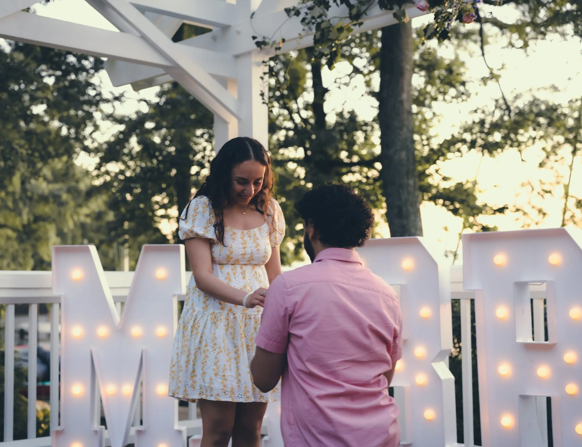 Marina looking down at George kneeling with the ring beside the marquee letters