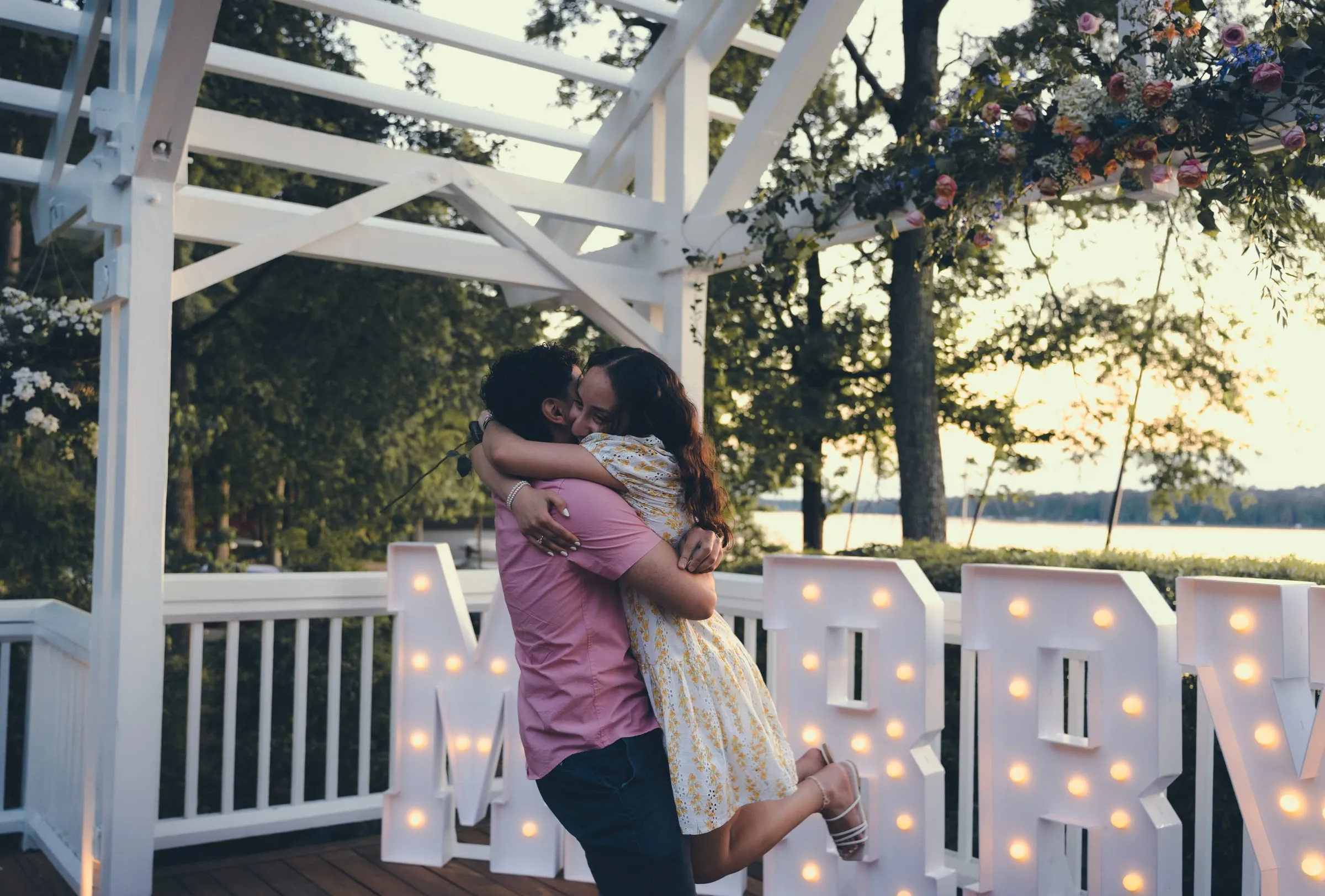George lifting Marina off the ground in a hug beneath the flower-draped pavilion