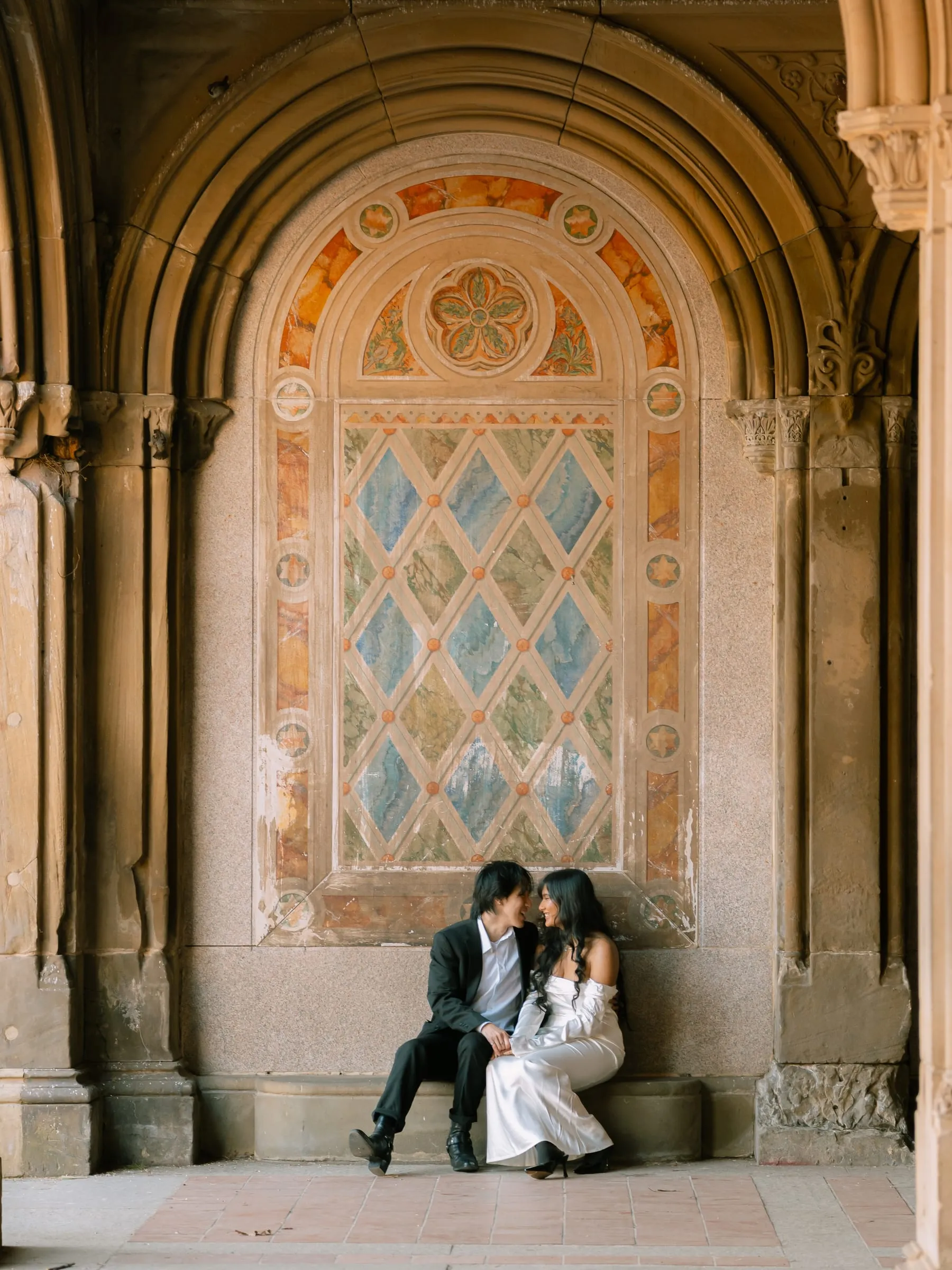 Noushin and Nathan smiling at each other beneath Bethesda Terrace's painted stone lattice panel