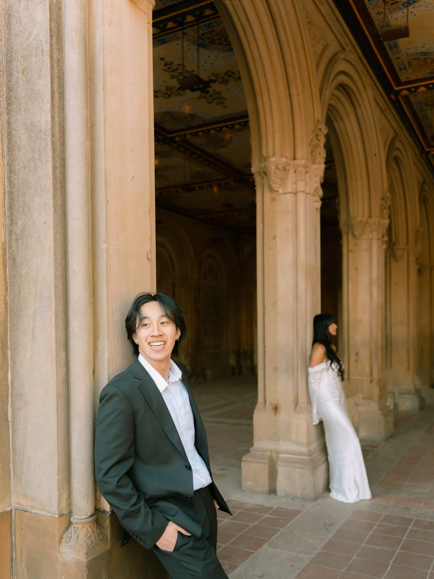 Nathan leaning against a column and smiling as Noushin waits by a column behind him
