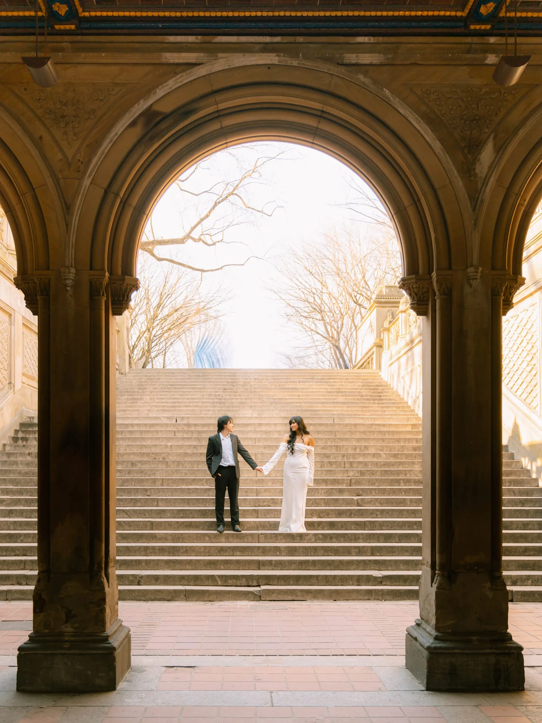 Noushin and Nathan holding hands on the grand staircase, framed by the stone arch below