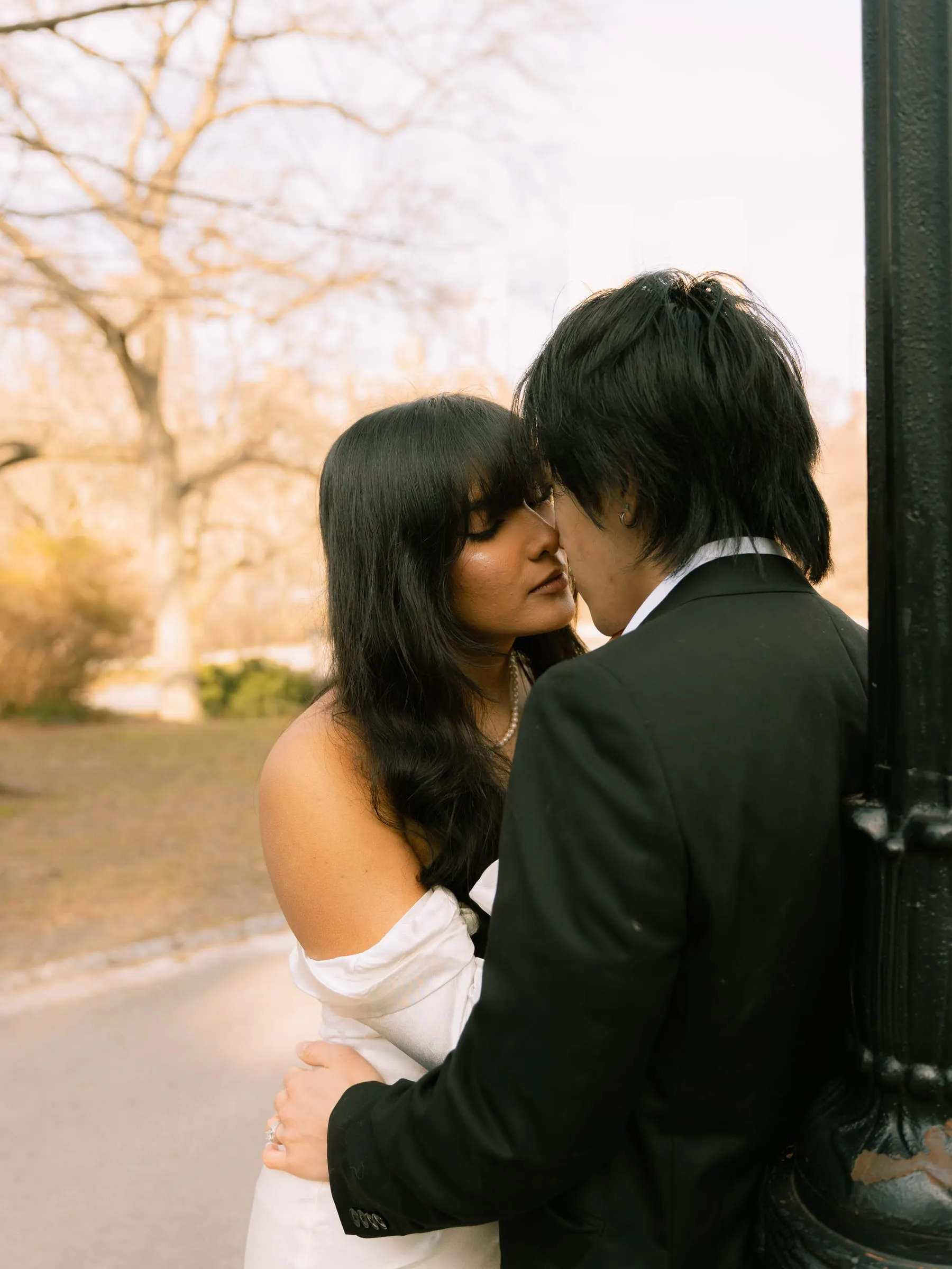 Noushin and Nathan nose to nose beside a lamppost, golden light through the bare trees behind them