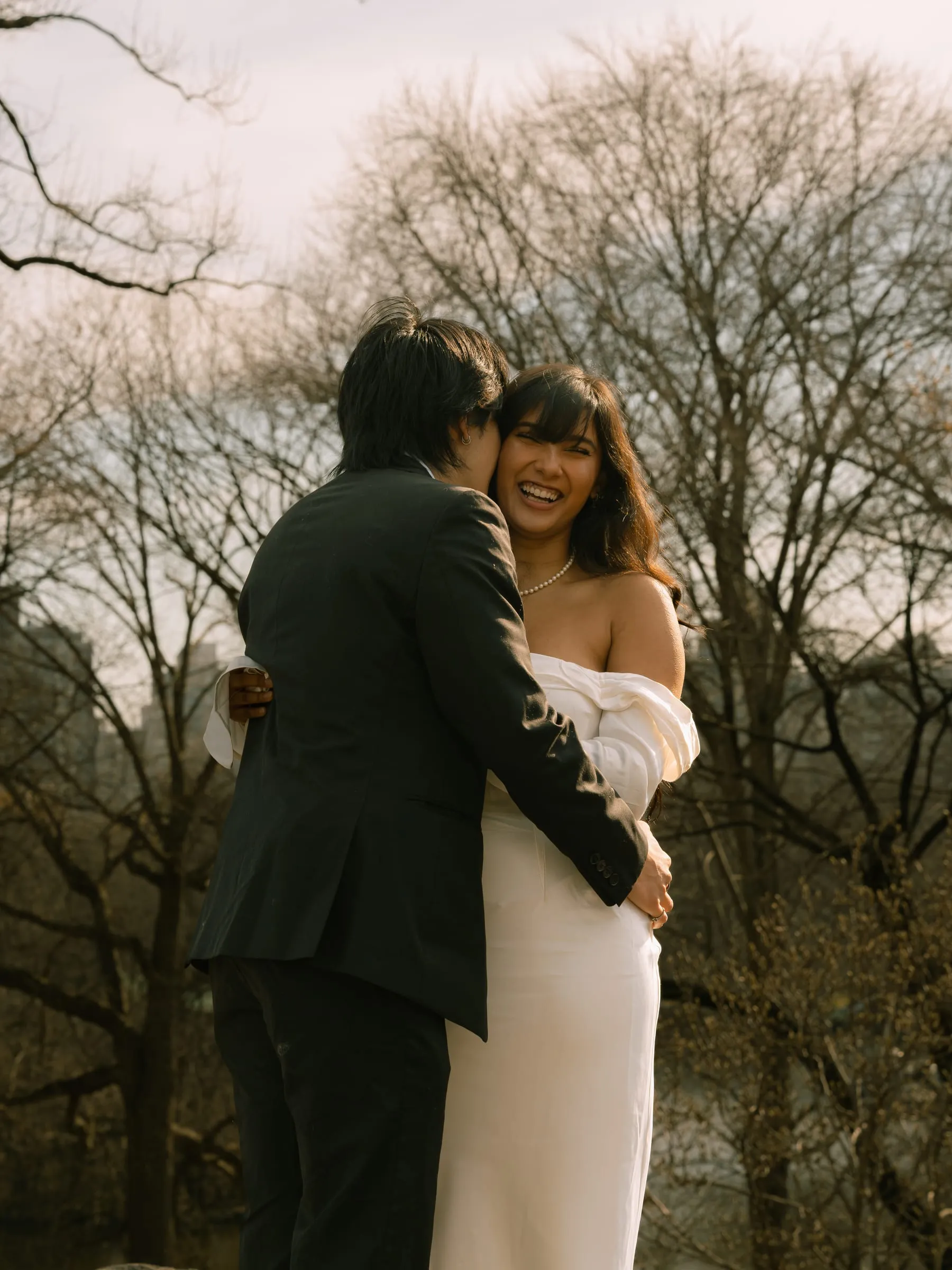 Nathan kissing Noushin's cheek as she laughs, the bare trees rising behind them