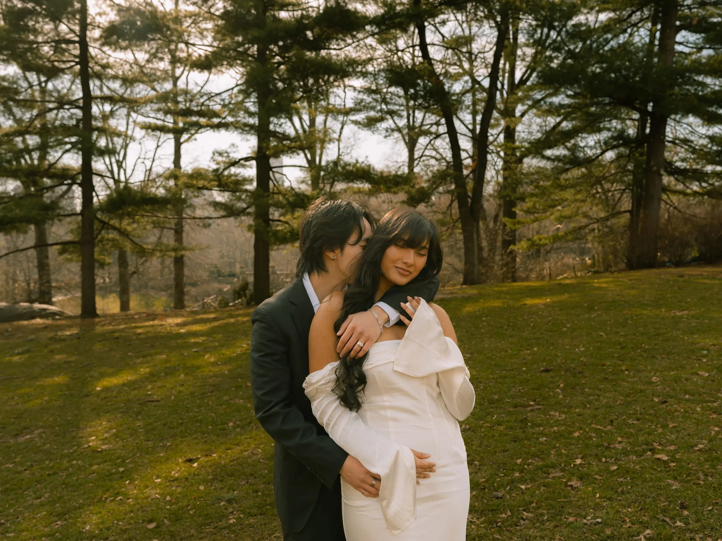 Nathan embracing Noushin from behind in a sunlit clearing among the pines