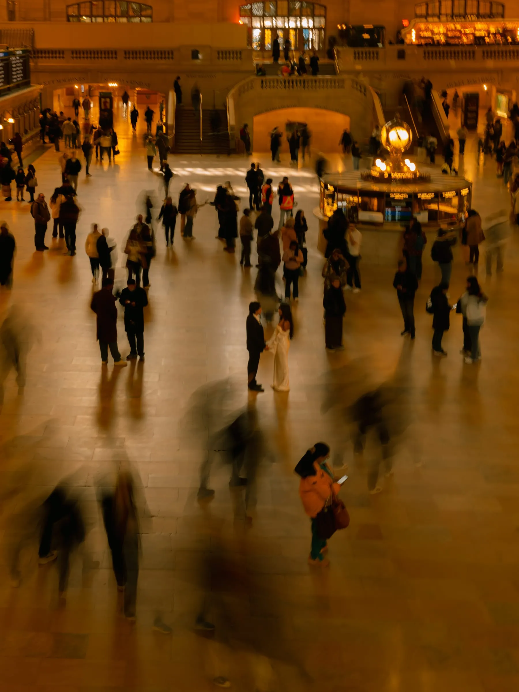 A wide view of Noushin and Nathan pausing together in Grand Central Terminal as travelers move around them