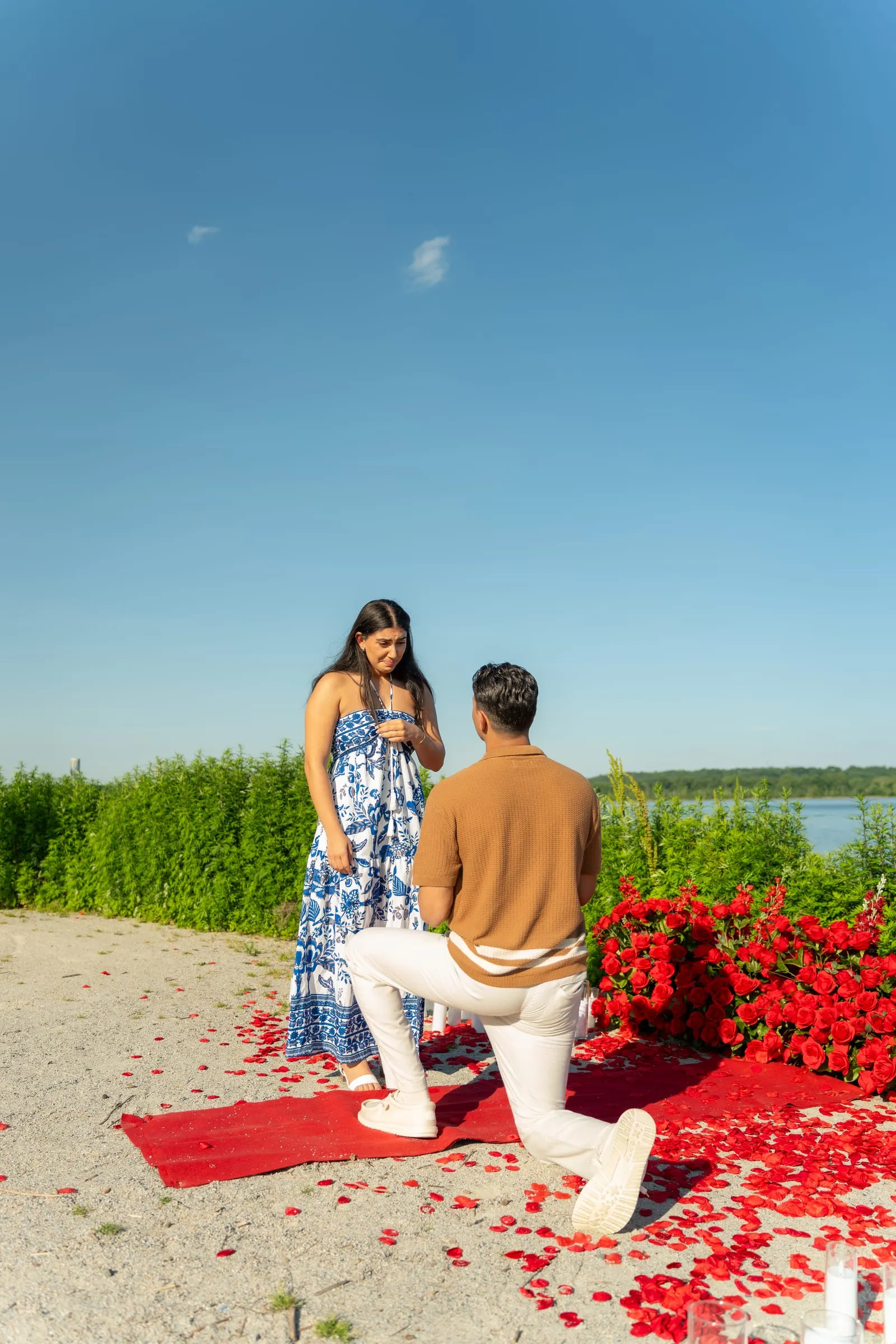 A wide view of Thomas kneeling among red roses and candles to propose to Verina