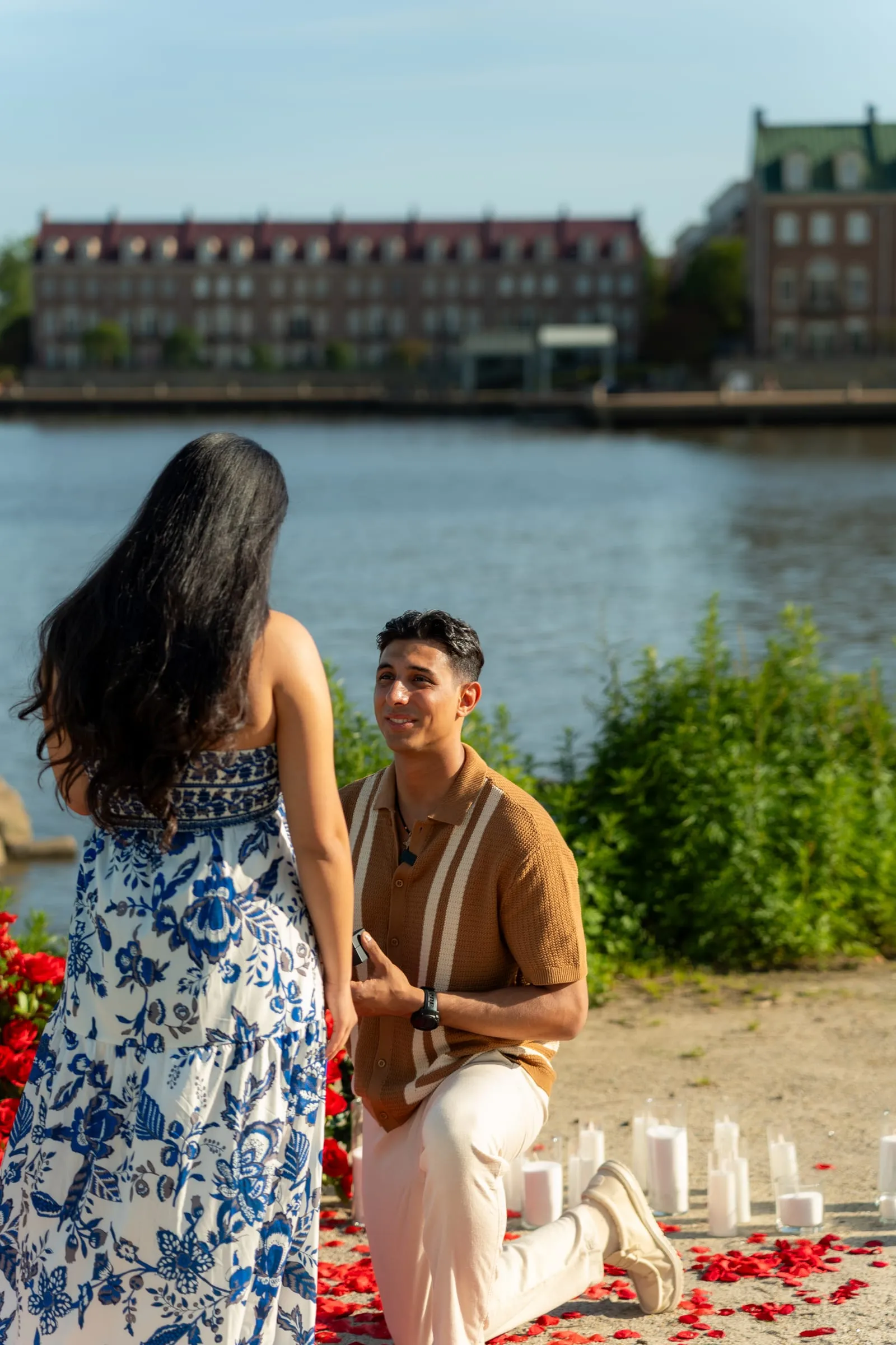 Thomas smiling up at Verina from one knee, the Old Town Alexandria waterfront behind them