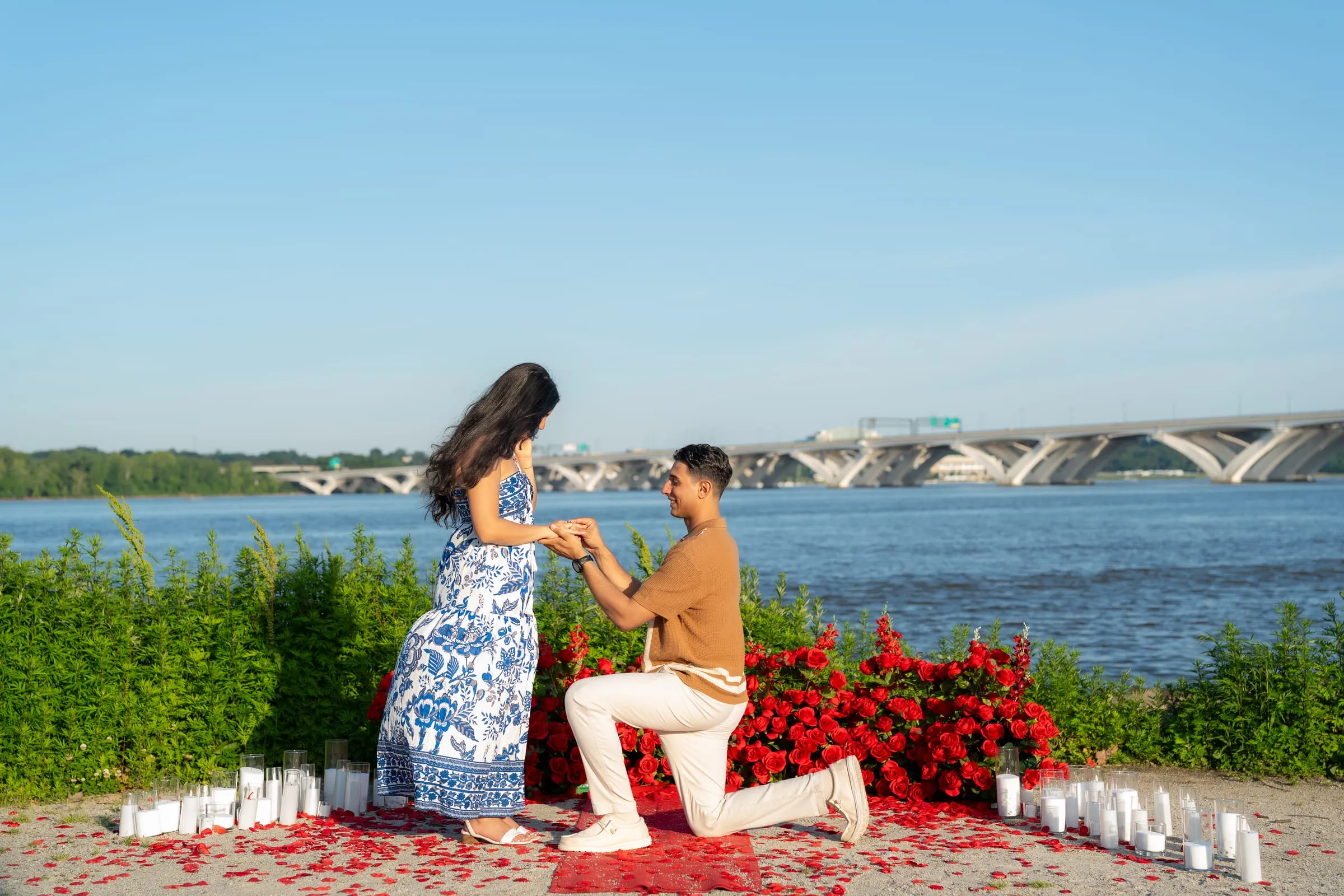 Thomas on one knee holding Verina's hand among the roses and candles, the Woodrow Wilson Bridge behind them